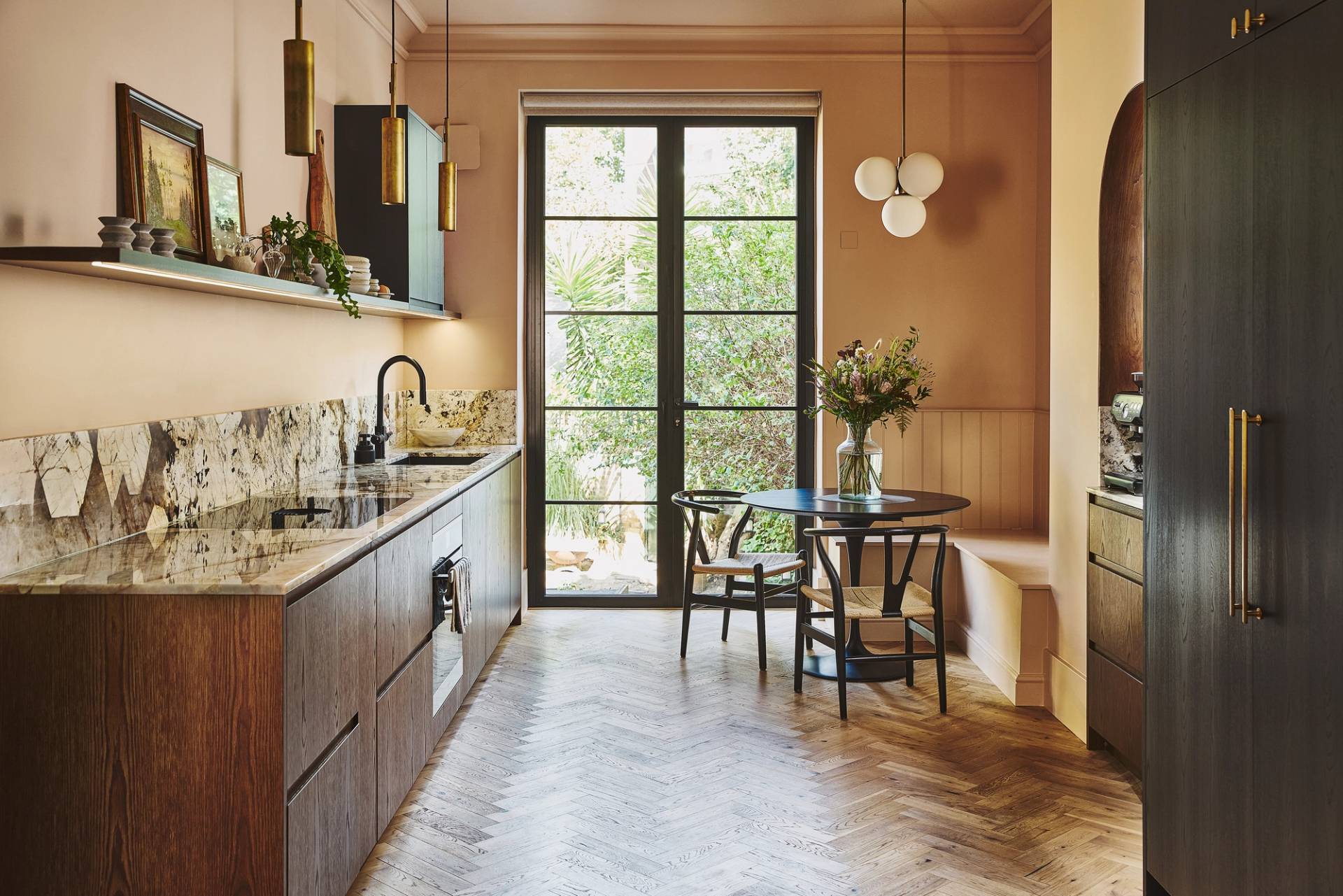 A modern kitchen with dark wood cupboards, stone worktops, a herringbone floor, open shelves, large windowed doors to a garden, a round table with chairs, and pendant lights. Flowers are arranged on the table