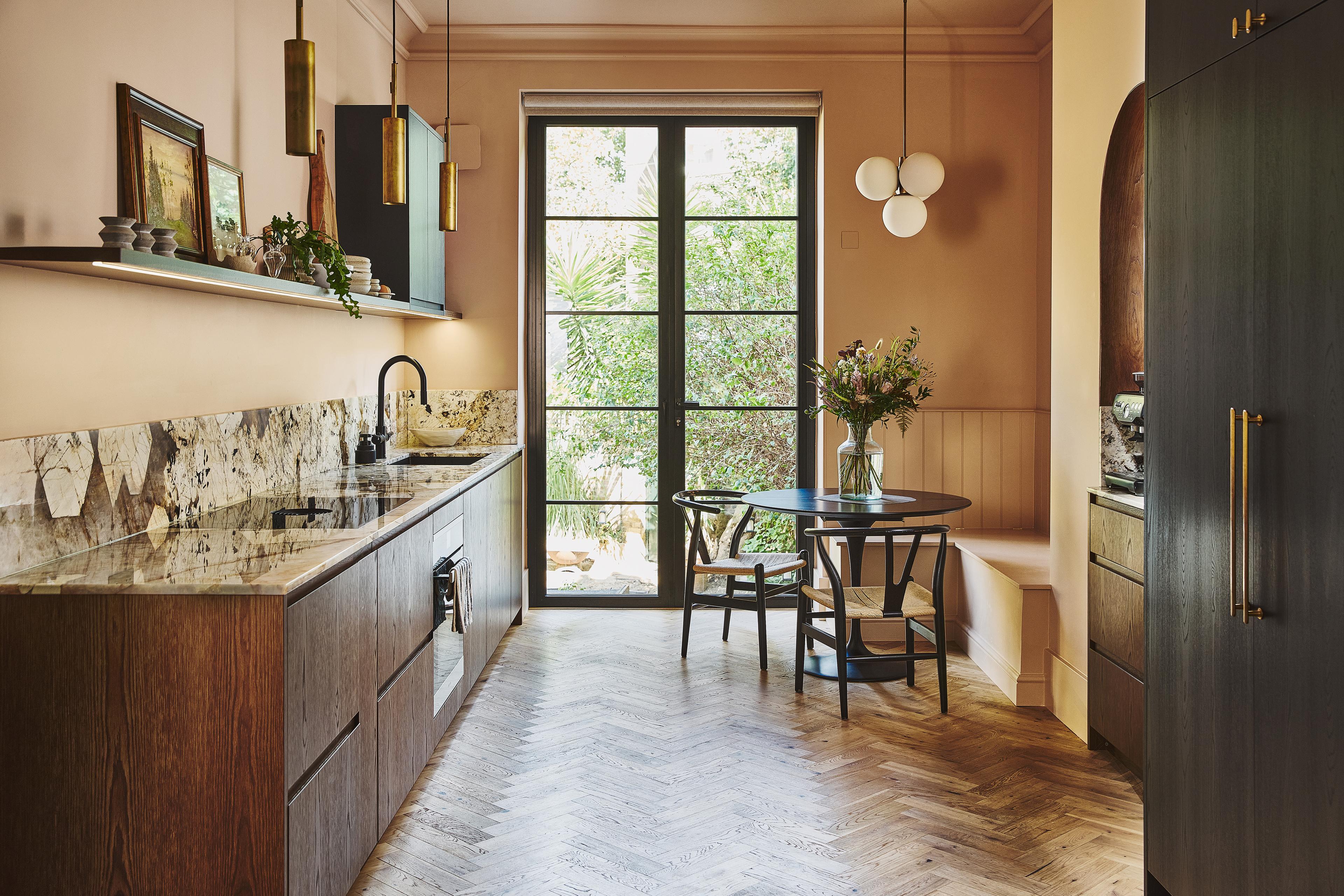 A modern kitchen with dark wood cupboards, stone worktops, a herringbone floor, open shelves, large windowed doors to a garden, a round table with chairs, and pendant lights. Flowers are arranged on the table