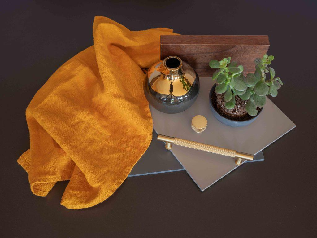 Top down shot of Interior design mood board flatlay featuring grey finish samples, knurled brass handles, a small metallic ceramic vase, a potted jade plant, and a draped mustard yellow linen cloth against a wood grain background.