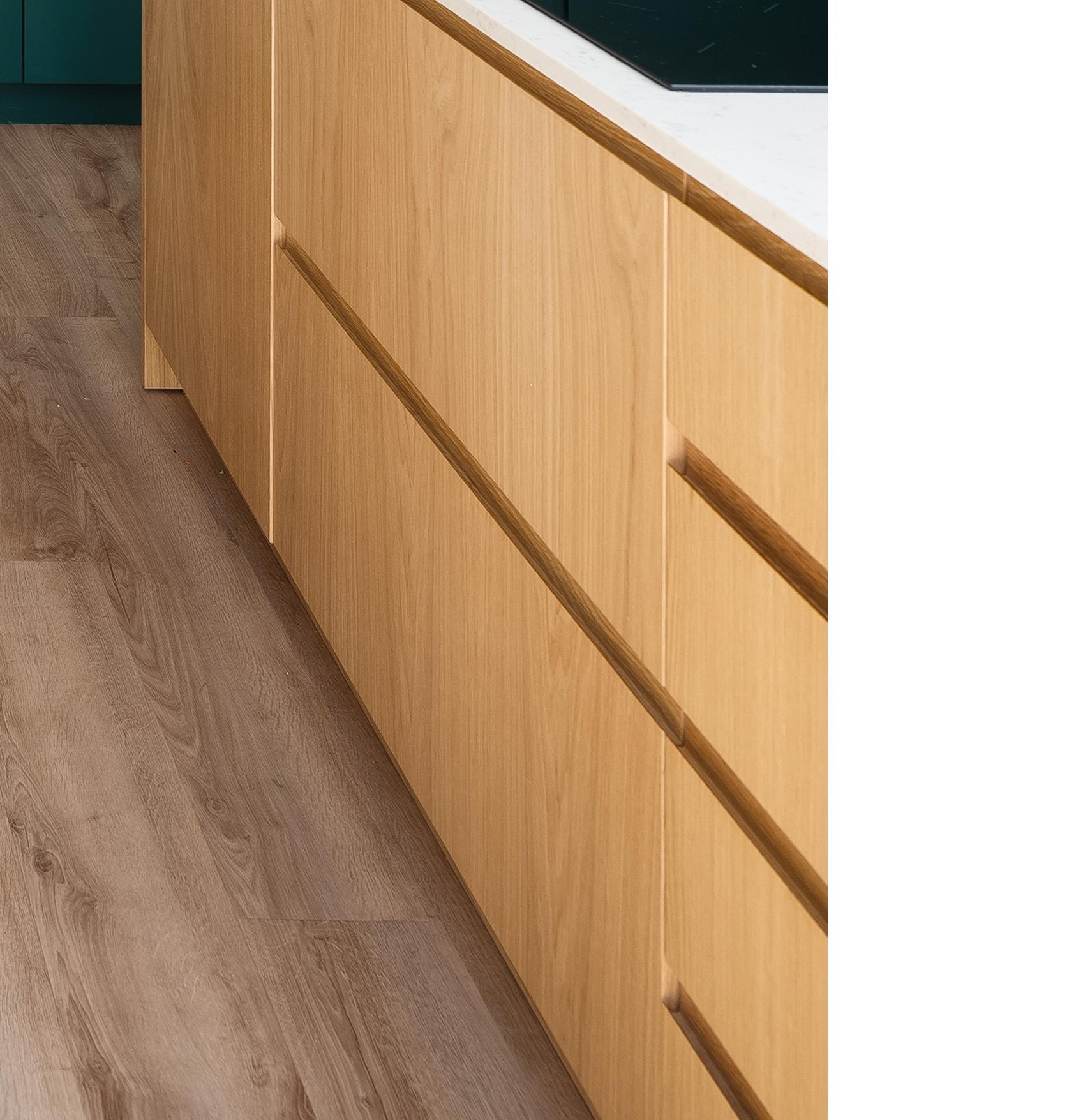 Light wood kitchen cabinets with flat fronts and integrated handles are shown next to a brown wood floor. A white countertop is partially visible at the top of the image.
