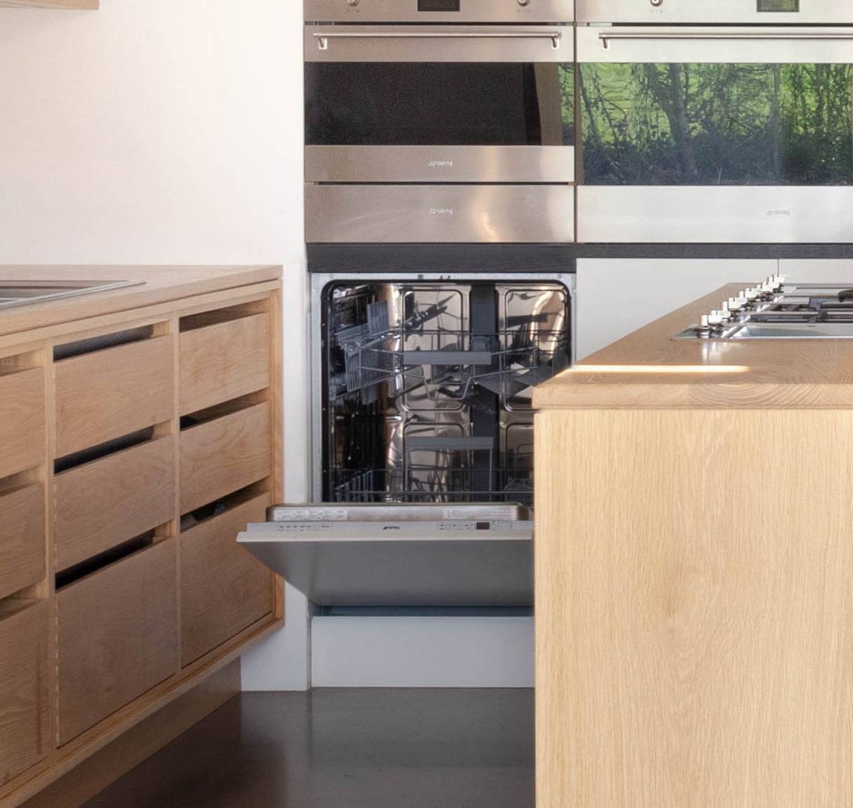A modern kitchen with a wooden finish. An open dishwasher is built into the cabinetry. Above it, there is a reflective oven. The countertop is made of wood, and a stovetop is installed on an island in the foreground.