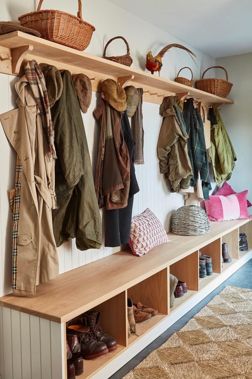 A cozy mudroom with a wooden bench and cubbies filled with shoes. Above, a shelf holds wicker baskets and hats. Various coats and jackets hang on hooks, and colorful cushions rest on the bench. A woven rug covers the dark floor.