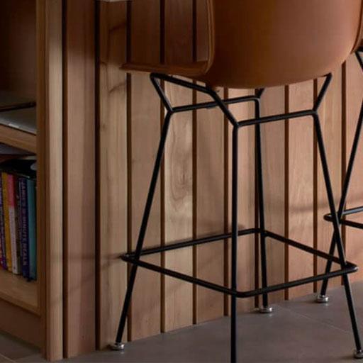 A close-up of a modern bar stool with a brown seat and black metal legs next to a wooden wall and a built-in bookshelf partially filled with books.