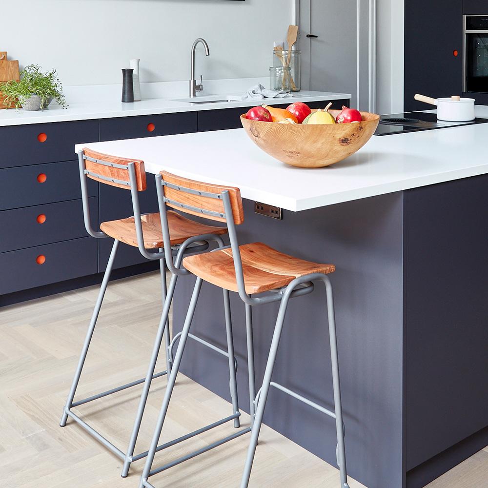 A modern kitchen with a white countertop island, two wooden bar stools with metal legs, a wooden fruit bowl filled with apples and bananas, dark cabinets with orange knobs, and a sink in the background.