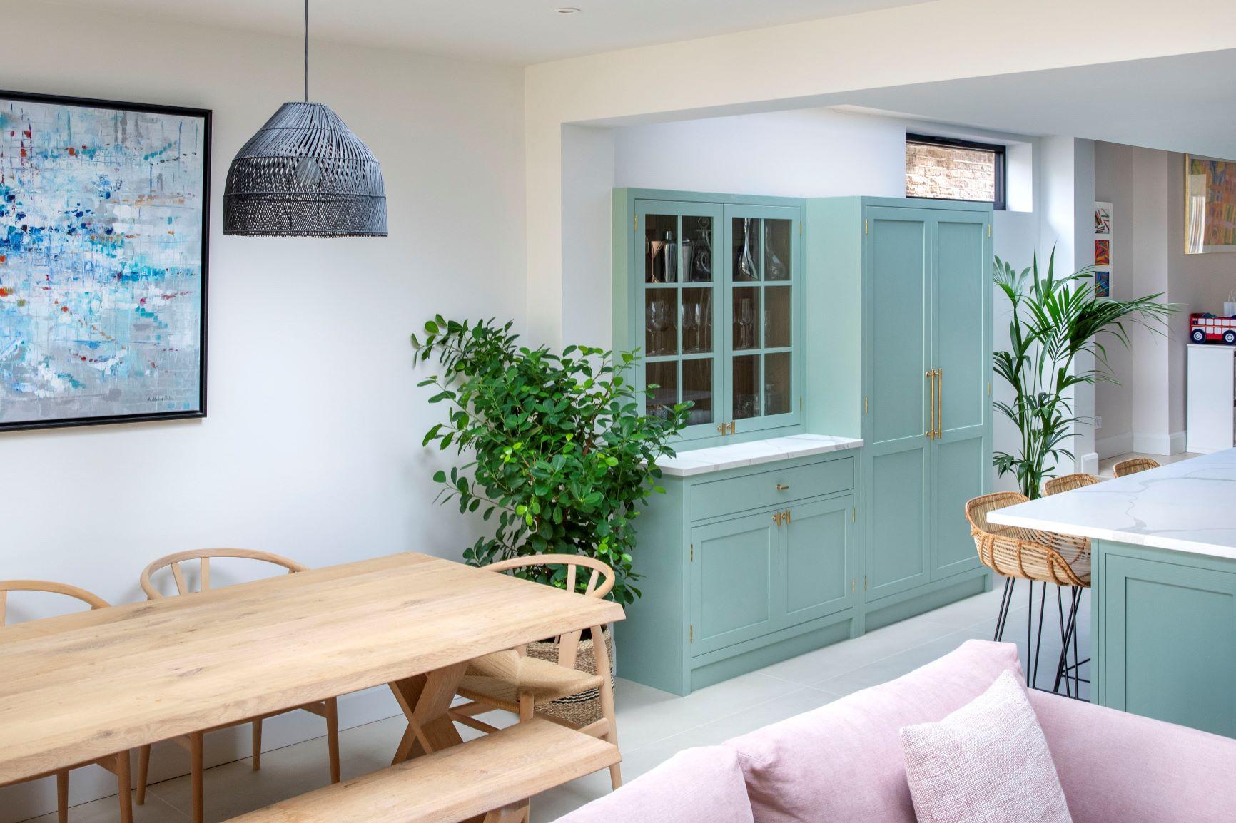 A captivating view of the Dulwich kitchen, featuring custom green cabinets, marble effect quartz worktops, and brass accents. The light green cabinets blend seamlessly with the pastel shades of the open-plan room. A generous island with ample countertop space enhances the functionality of the kitchen.