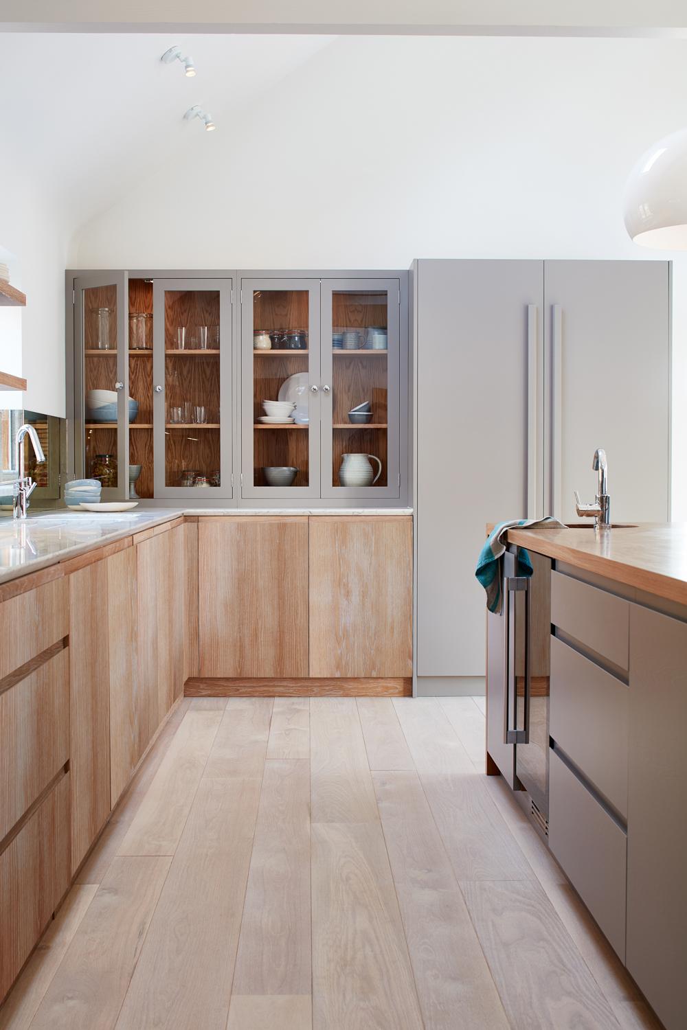 A modern kitchen features light wooden cabinets and flooring, open shelves with dishes, and a sleek, gray refrigerator. Glass-front cabinets display neatly arranged tableware. A kitchen island with drawers and a sink is adorned with a turquoise towel.