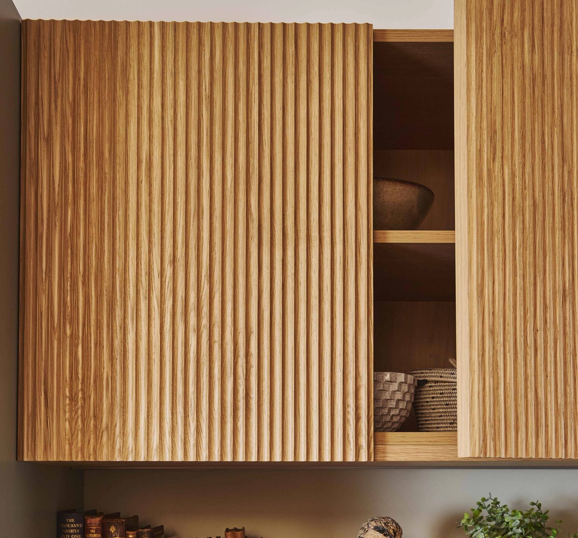 A modern kitchen features a wooden island with a black metal bar stool, a white countertop, a stove, green cabinets, and a small potted plant by a sunlit window.