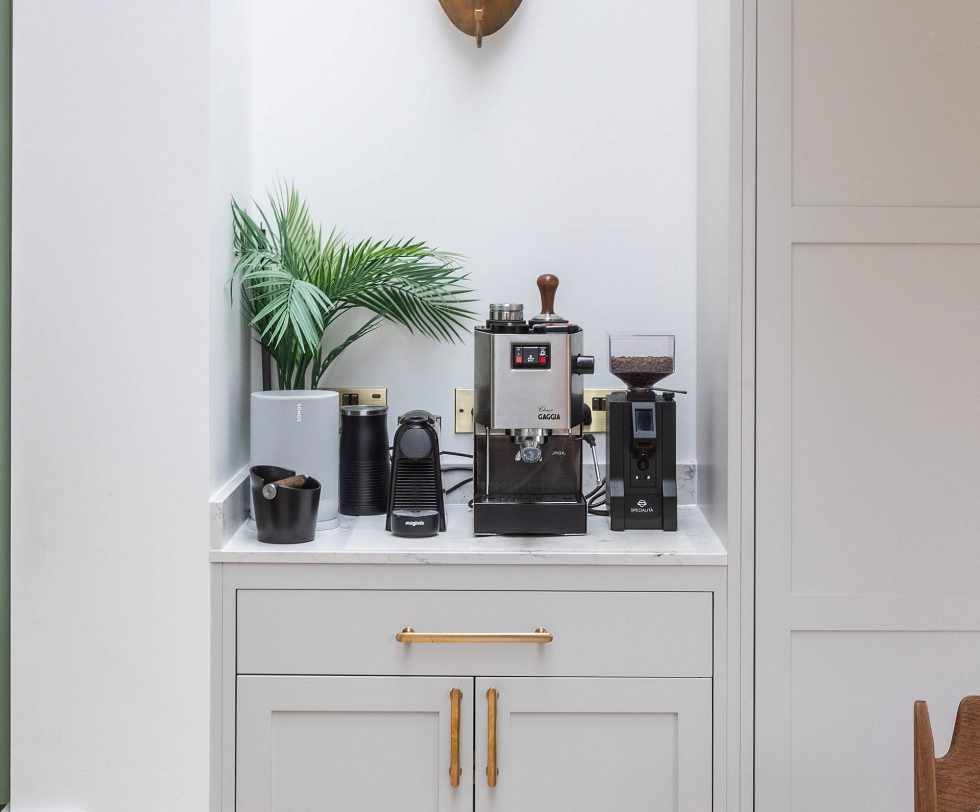 A small coffee station with a coffee machine, grinder, and accessories sits on a white marble countertop, with a potted green plant and two black canisters in the corner, above light gray cabinets with gold handles.