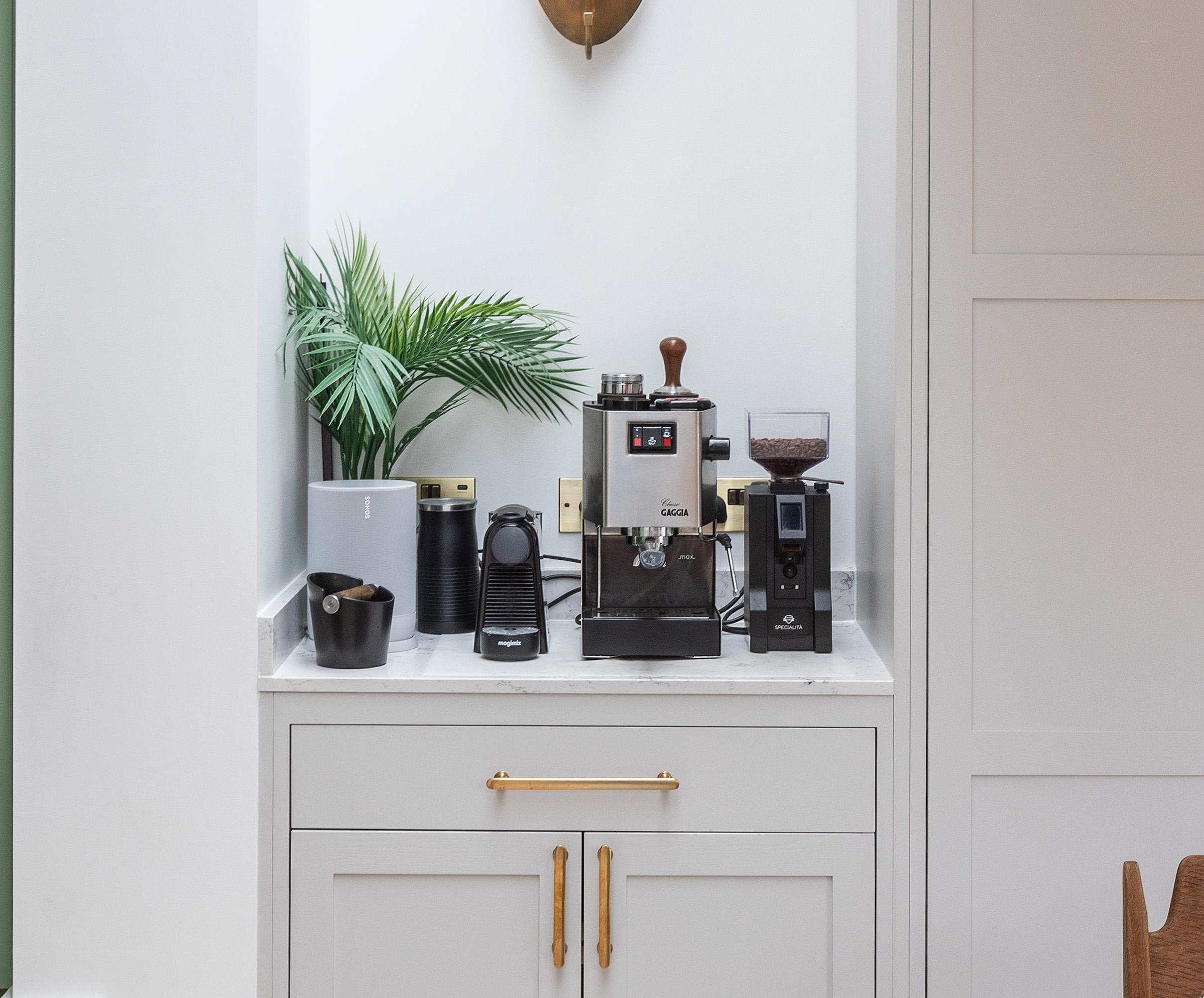 A small coffee station with a coffee machine, grinder, and accessories sits on a white marble countertop, with a potted green plant and two black canisters in the corner, above light gray cabinets with gold handles.