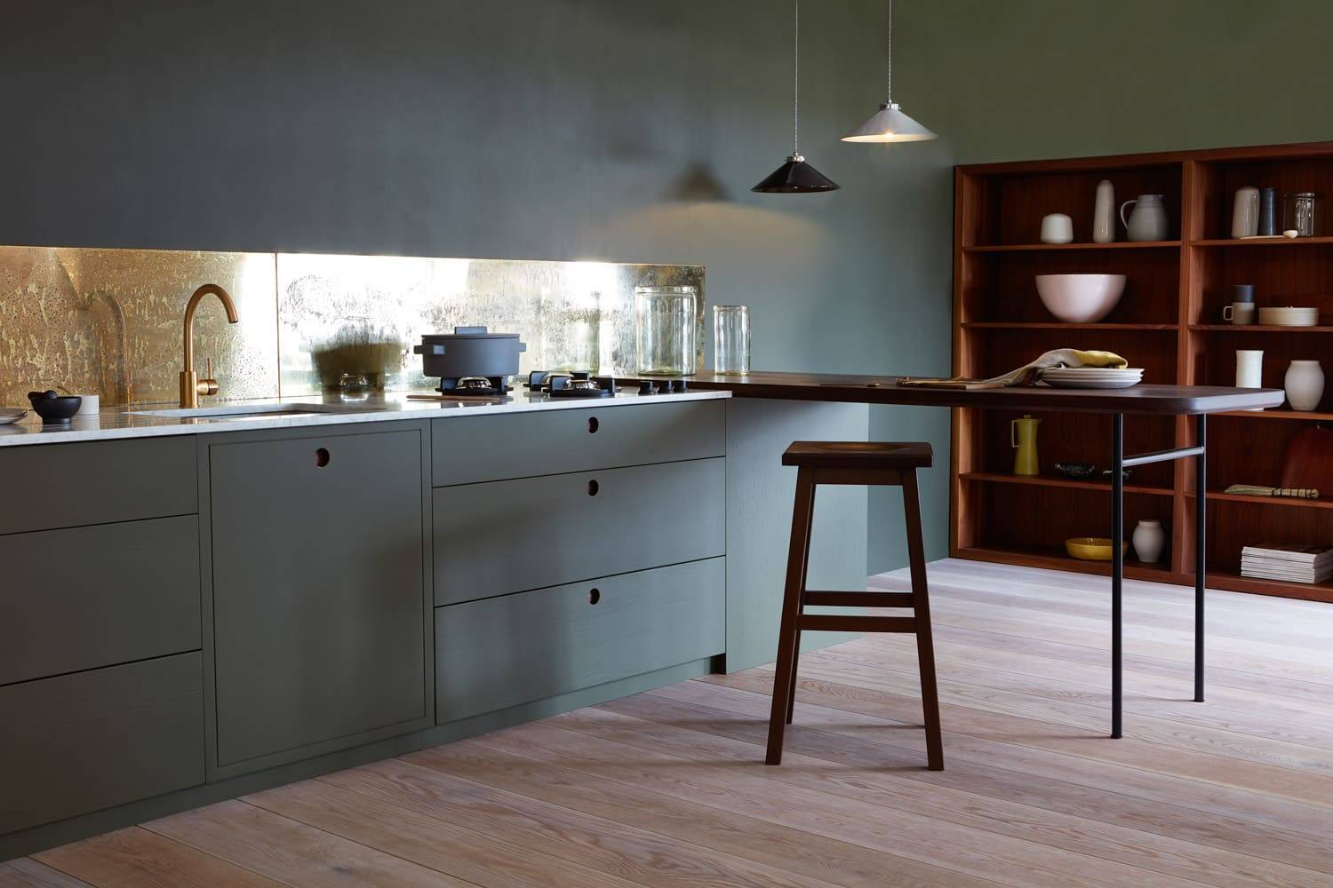 Modern kitchen with green cabinets, a wood countertop, and a tall stool. A metallic backsplash reflects light above the sink. Shelves display dishes, bowls, and vases. Pendant lights hang from the ceiling, and the floor is light wood.