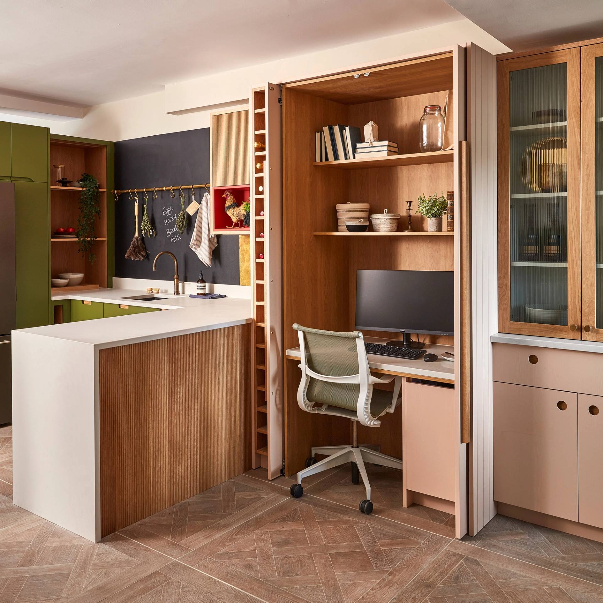Modern kitchen with wood accents and green cabinets, featuring a hidden home office nook with a desk, shelves, computer, and office chair tucked behind folding doors. The space is tidy and well-organized.