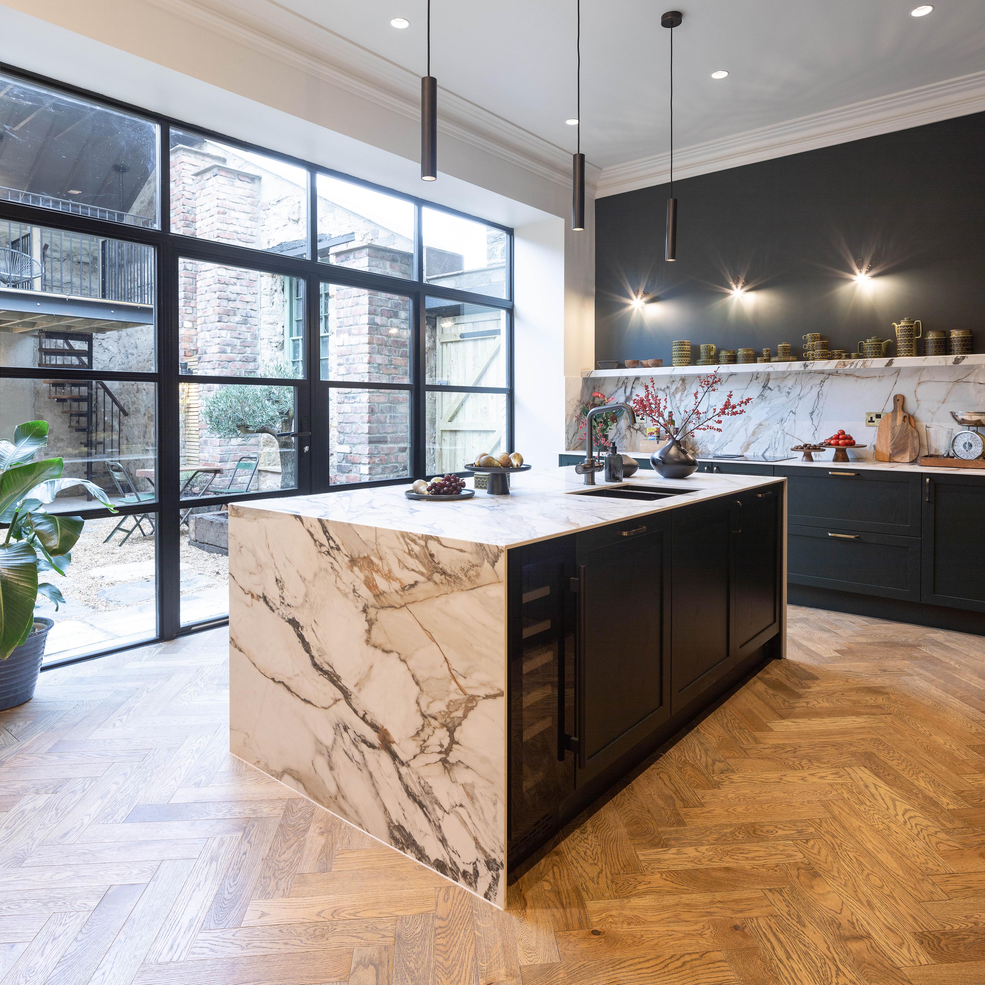 Modern kitchen featuring a large marble island with black cabinets and a sink. The room has dark walls, wooden herringbone flooring, and large glass doors leading outside. Pendant lights hang above the island, and shelves display decorative items.