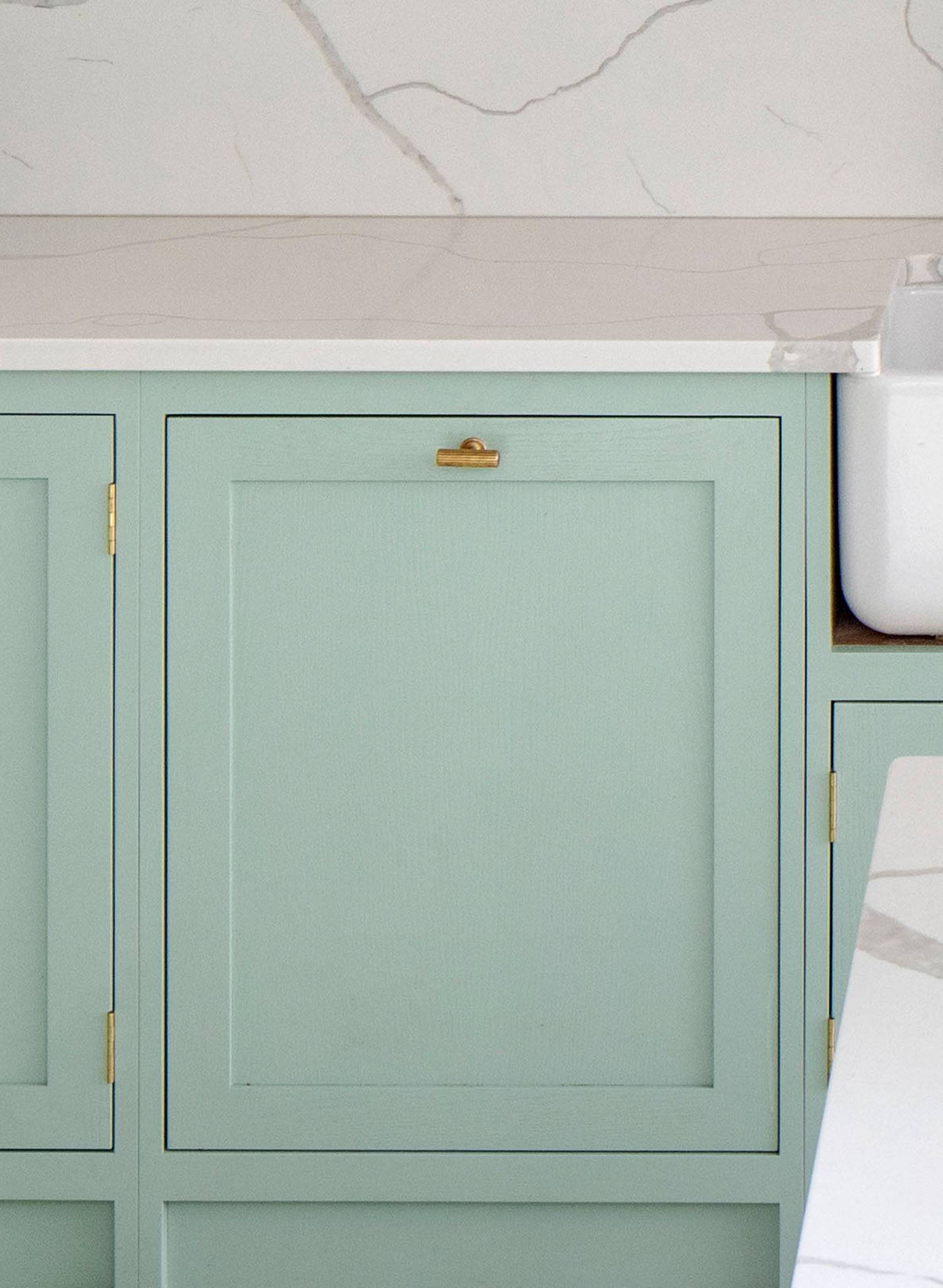 A pale green kitchen cabinet with a brass handle and hinges sits beneath a white marble countertop with gray veining; part of a white sink is visible on the right.