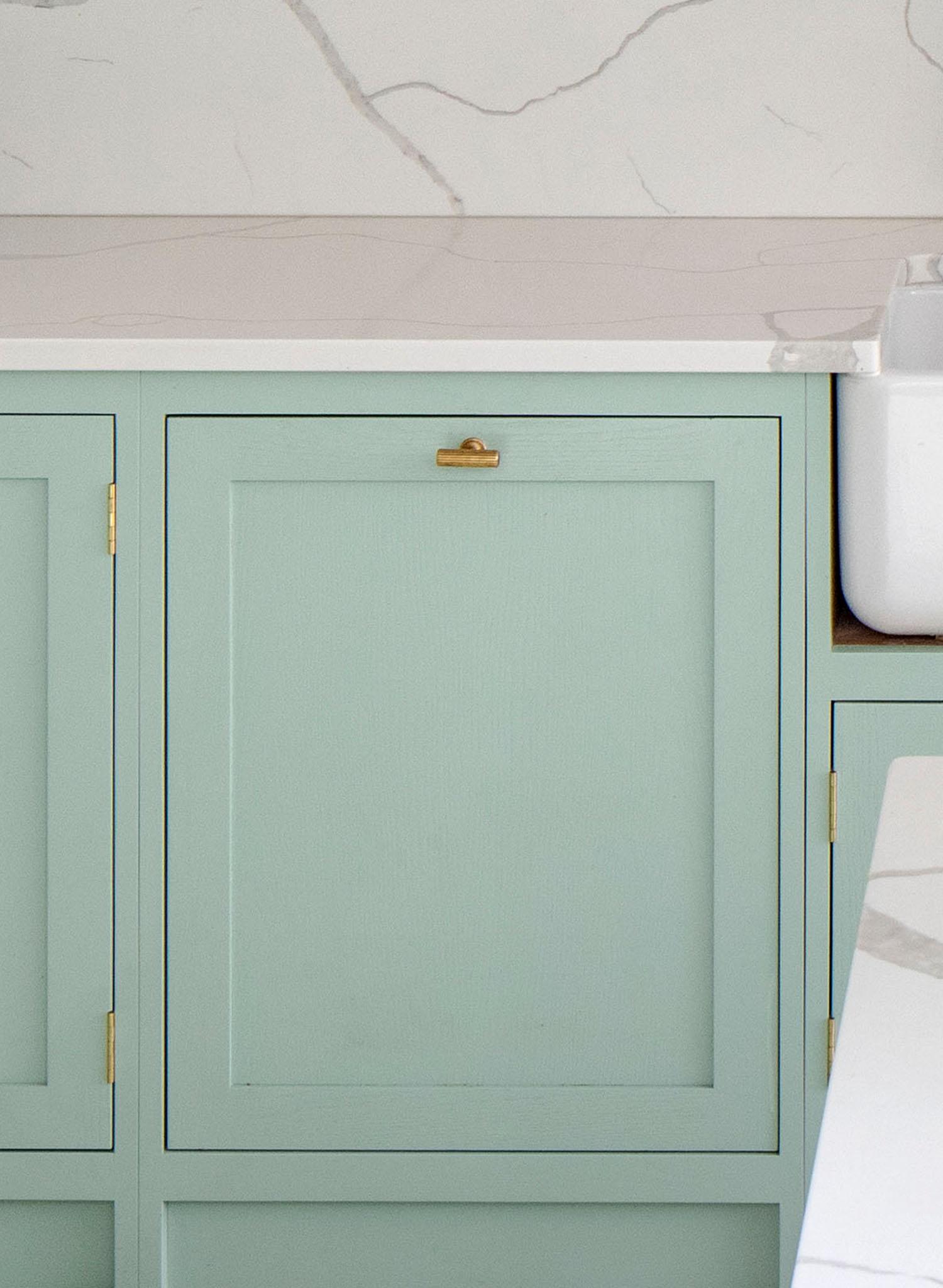 A pale green kitchen cabinet with a brass handle and hinges sits beneath a white marble countertop with gray veining; part of a white sink is visible on the right.