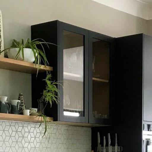 A kitchen with a dark glass-front cabinet, a wooden shelf holding green potted plants, ceramic mugs, and a geometric tile backsplash. A knife block sits on the counter below