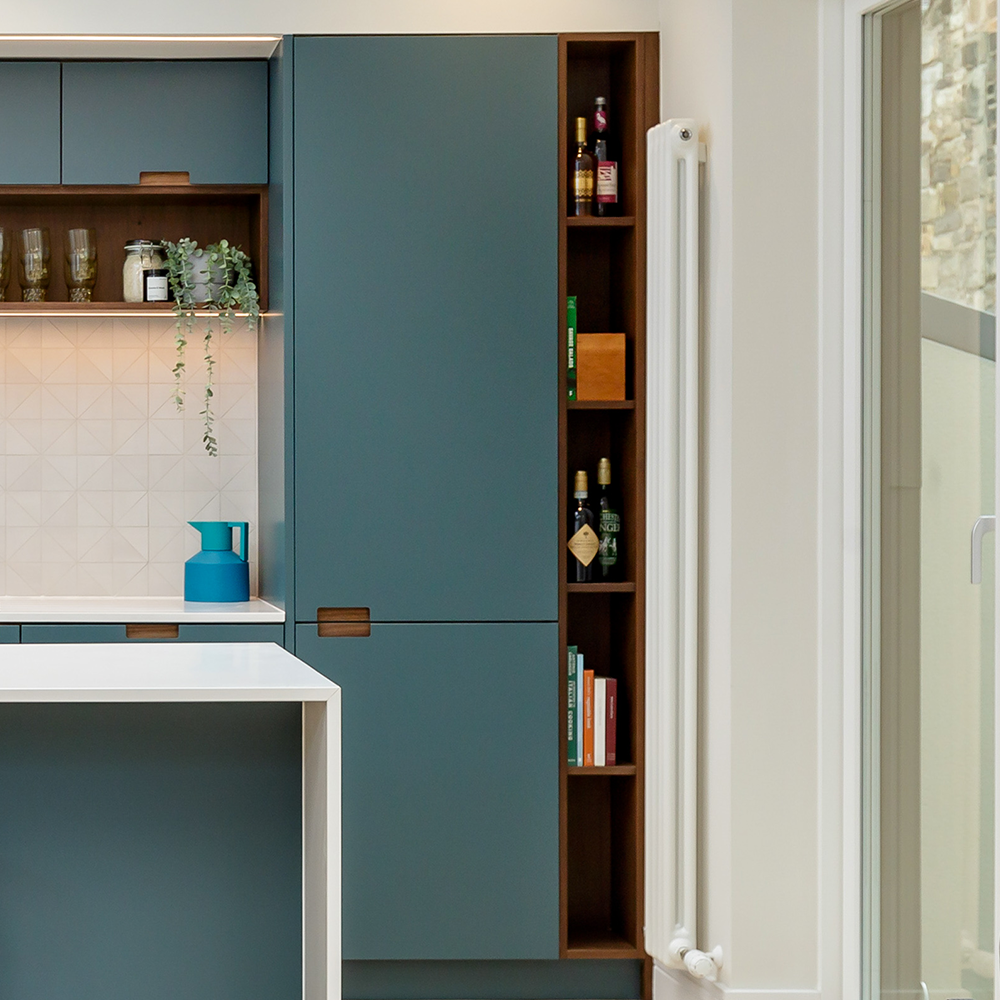 Modern kitchen with teal cabinets, a white countertop, and open shelves displaying glassware, bottles, and books. A blue pitcher sits on the counter, and a tall window brings in natural light.