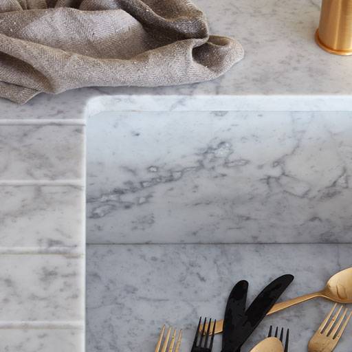 A marble kitchen sink with gold and black cutlery inside; a beige cloth rests on the marble counter, and a gold-colored utensil holder is partially visible on the right.