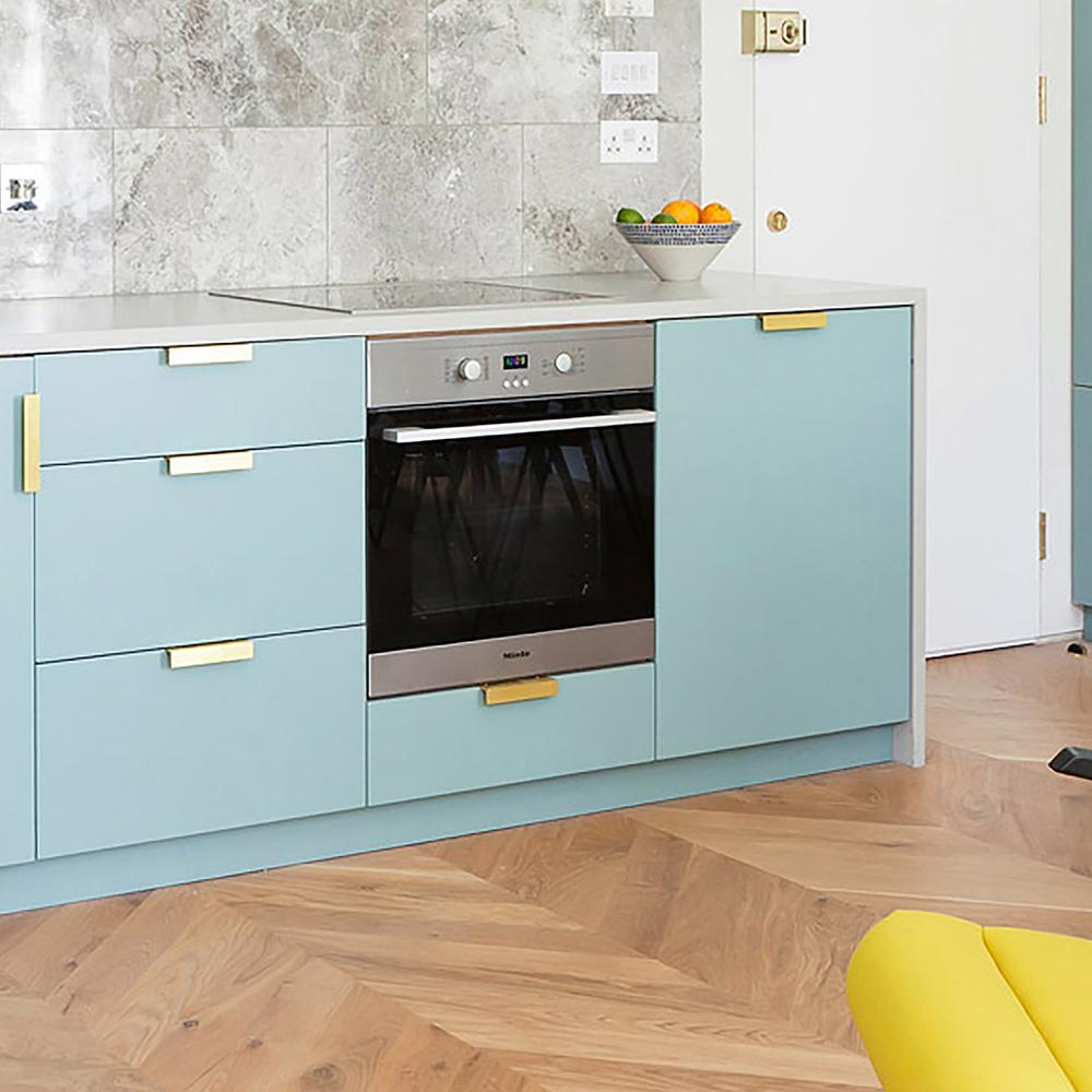 Modern light blue kitchen cabinets with brass pull handles, featuring a built-in stainless steel oven, white countertop, grey marble backsplash, and herringbone wood floors.