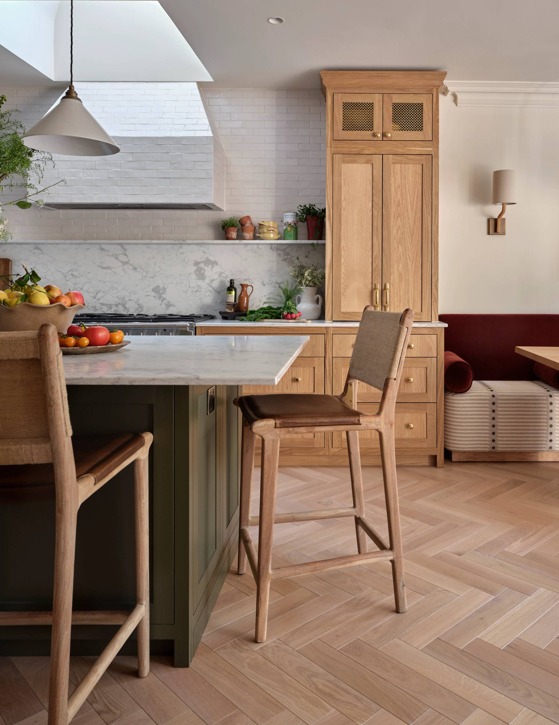 A modern kitchen with light wood cabinets, a marble countertop, bar stools, herringbone wood flooring, and a cozy built-in bench. Fruit and plants add color to the bright, inviting space.