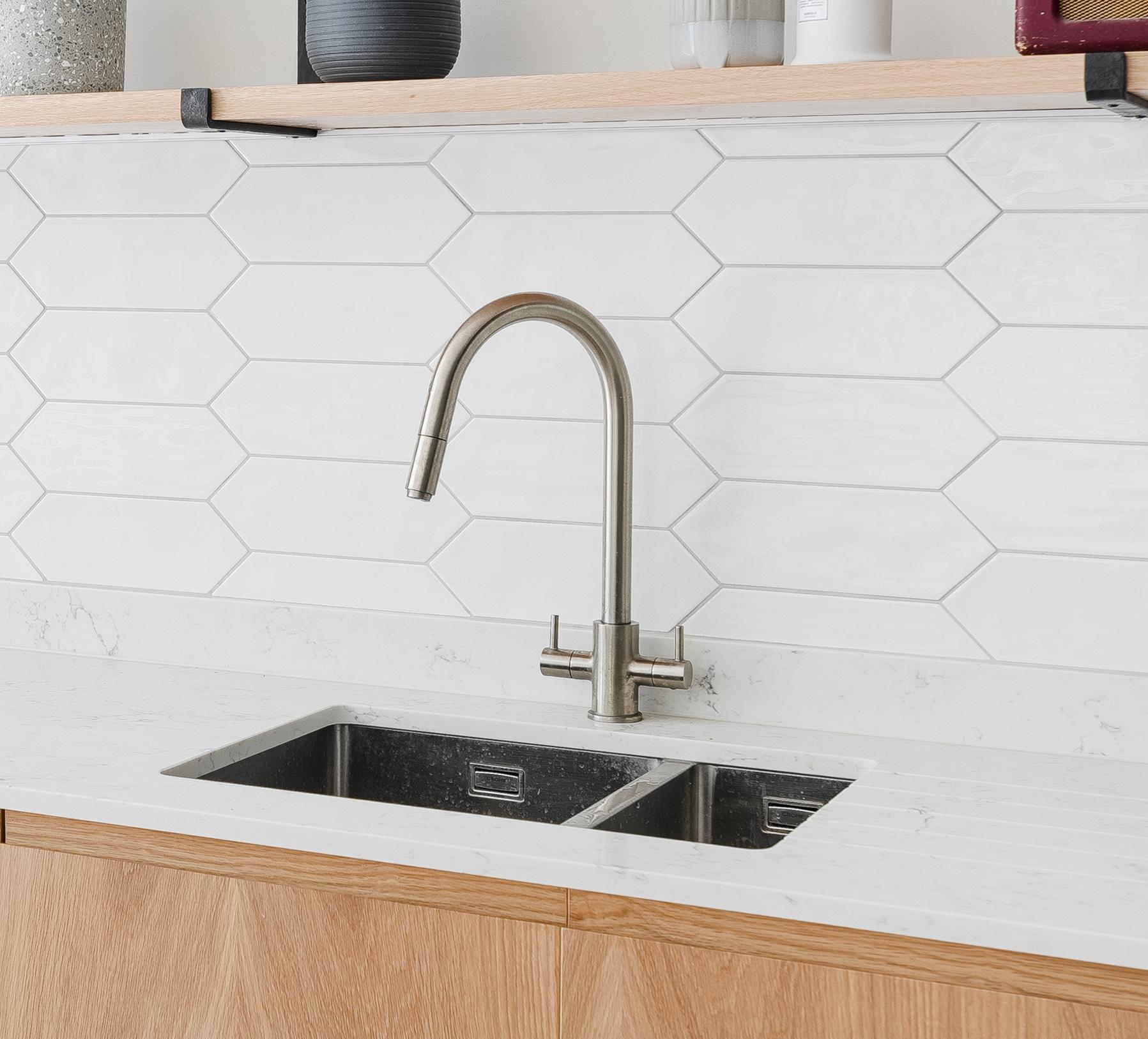 A modern kitchen sink with a stainless steel faucet, set in a white marble countertop. The backsplash features white hexagonal tiles, and there are light wood cabinets below.