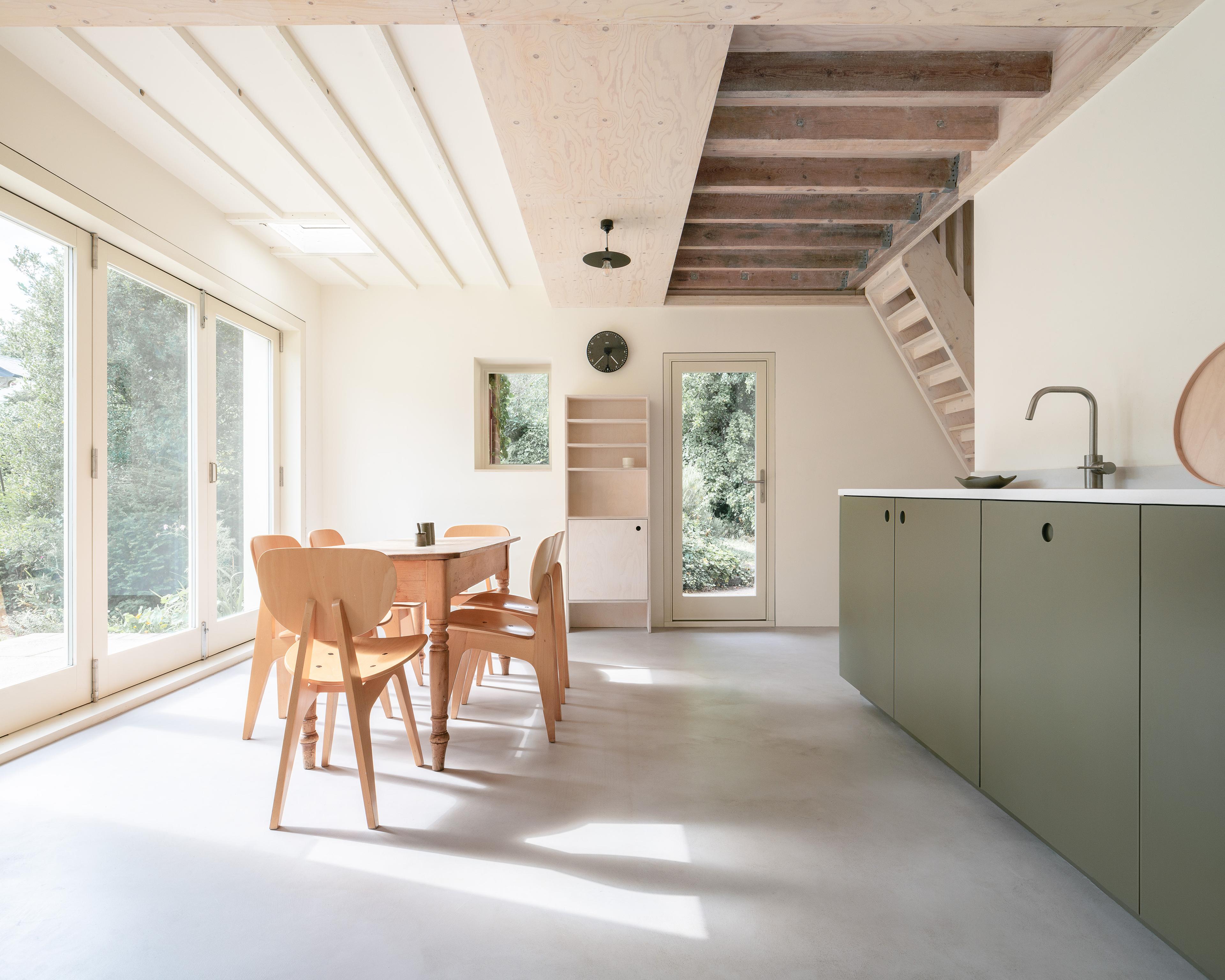 Minimalist kitchen and dining area with light wood furniture and green cabinetry. Large windows and glass doors on the left bring in natural light, overlooking greenery. Exposed beams are visible on the ceiling, and a wooden staircase leads upstairs.