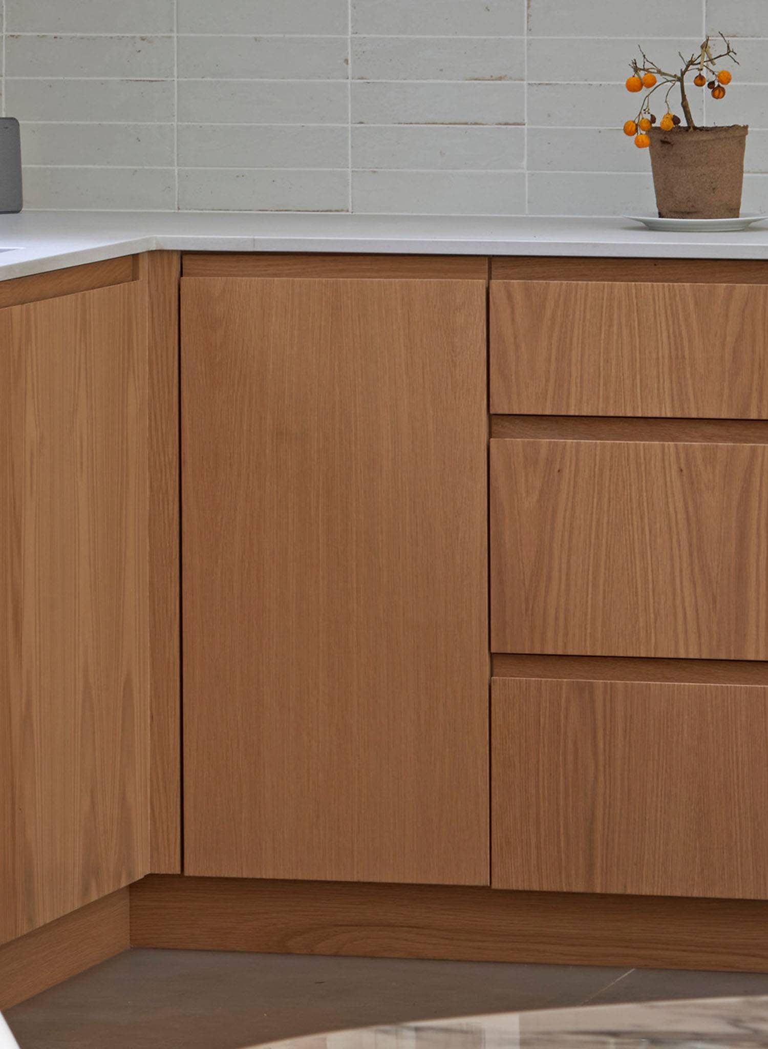 A minimalist kitchen corner with light wood cabinets, white countertop, and a small potted plant with orange berries against a white tiled backsplash.