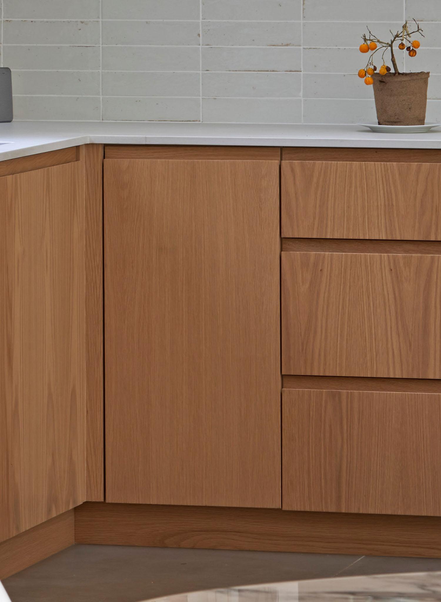 A minimalist kitchen corner with light wood cabinets, white countertop, and a small potted plant with orange berries against a white tiled backsplash.