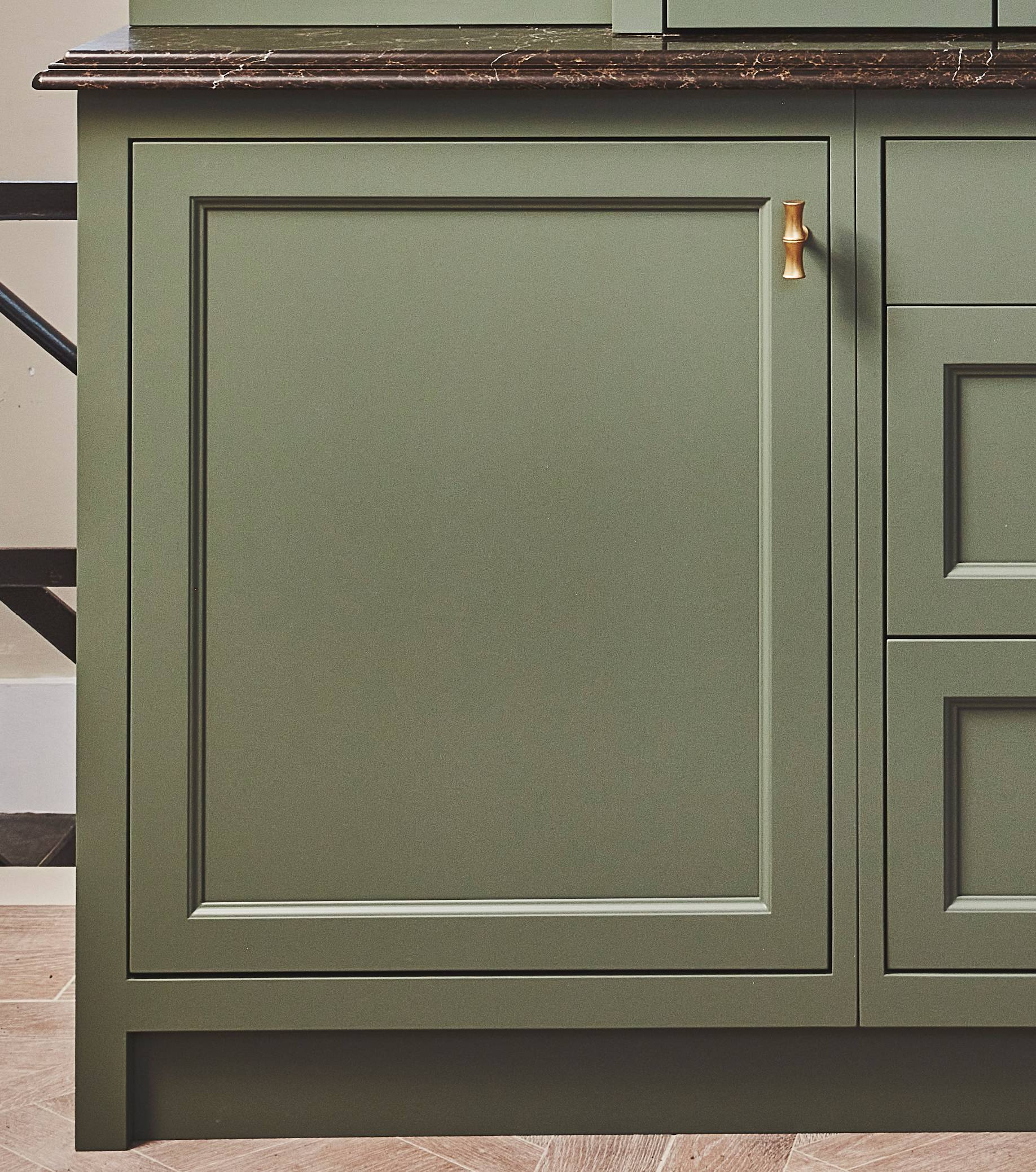 A green kitchen cabinet with a gold handle and a dark marble countertop, shown against a herringbone wood floor.