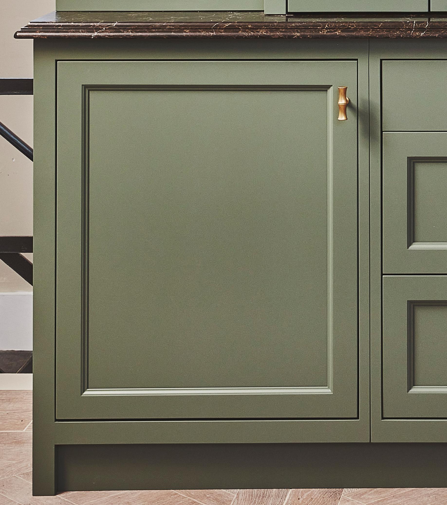 A green kitchen cabinet with a gold handle and a dark marble countertop, shown against a herringbone wood floor.