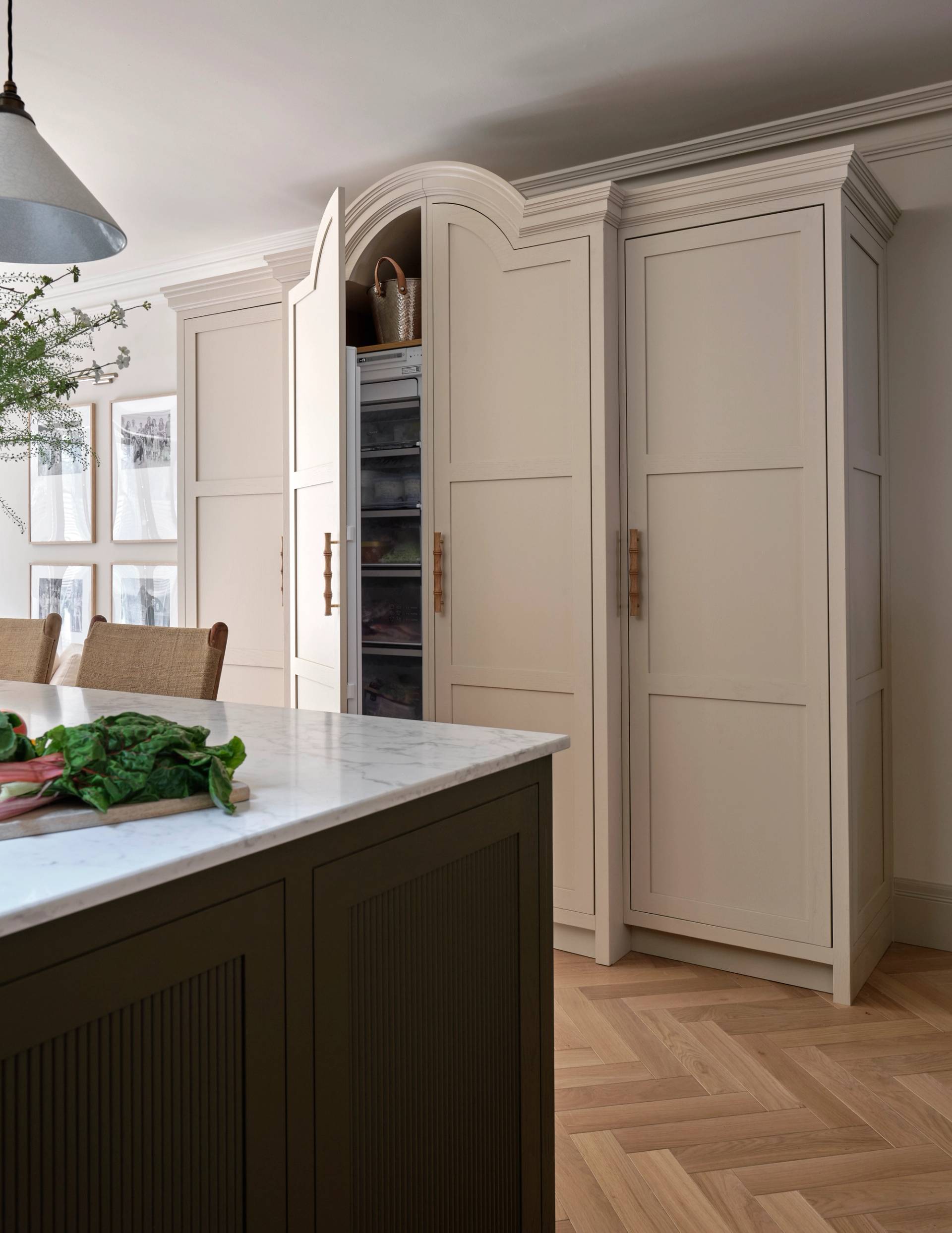 Large cream-colored kitchen cabinets with panel doors and brass handles stand against the wall. An island with a marble countertop and dark base is in the foreground. The floor features a light herringbone wood pattern.