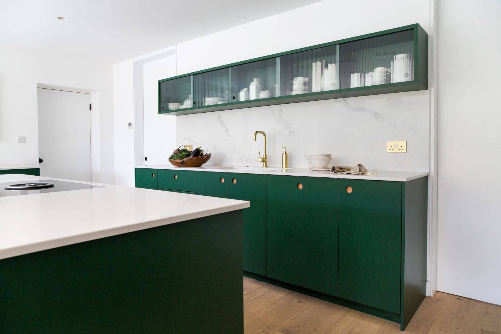 Modern kitchen with dark green cabinets, white countertops, a gold faucet, and open glass-fronted upper shelves displaying white dishes. The space has light wood flooring and a minimalist design.