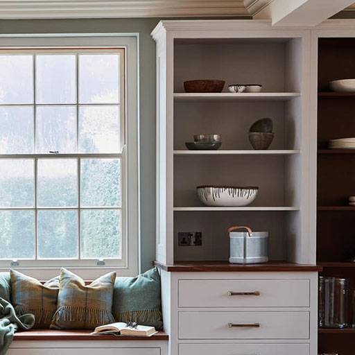 A cozy kitchen corner with a window seat adorned with green and beige pillows, beside white shelves displaying bowls and ceramics, and a countertop with a canister. Soft natural light fills the space.