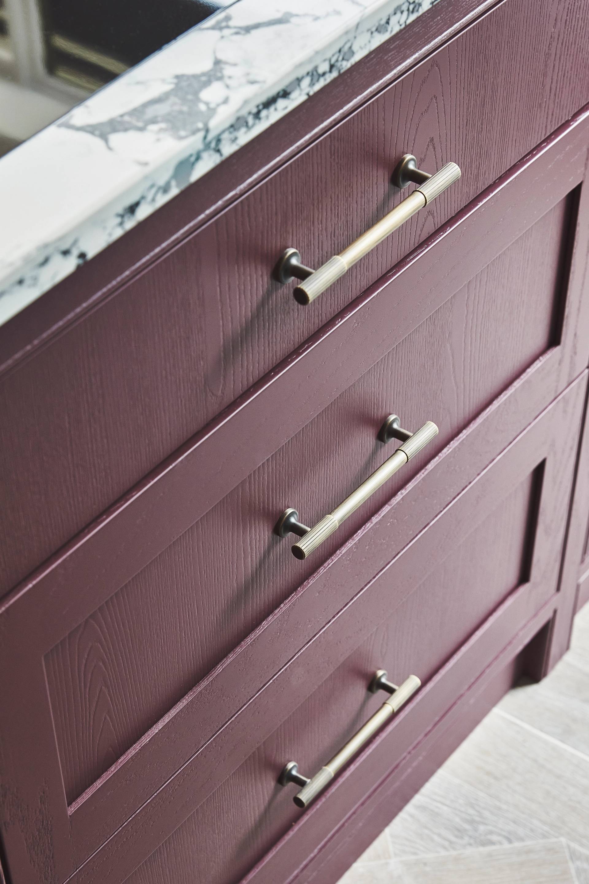 Close-up of a cabinet with three purple wooden drawers, each featuring a silver handle. The top is made of white marble with gray veining. The cabinet stands on a light-colored wooden floor.