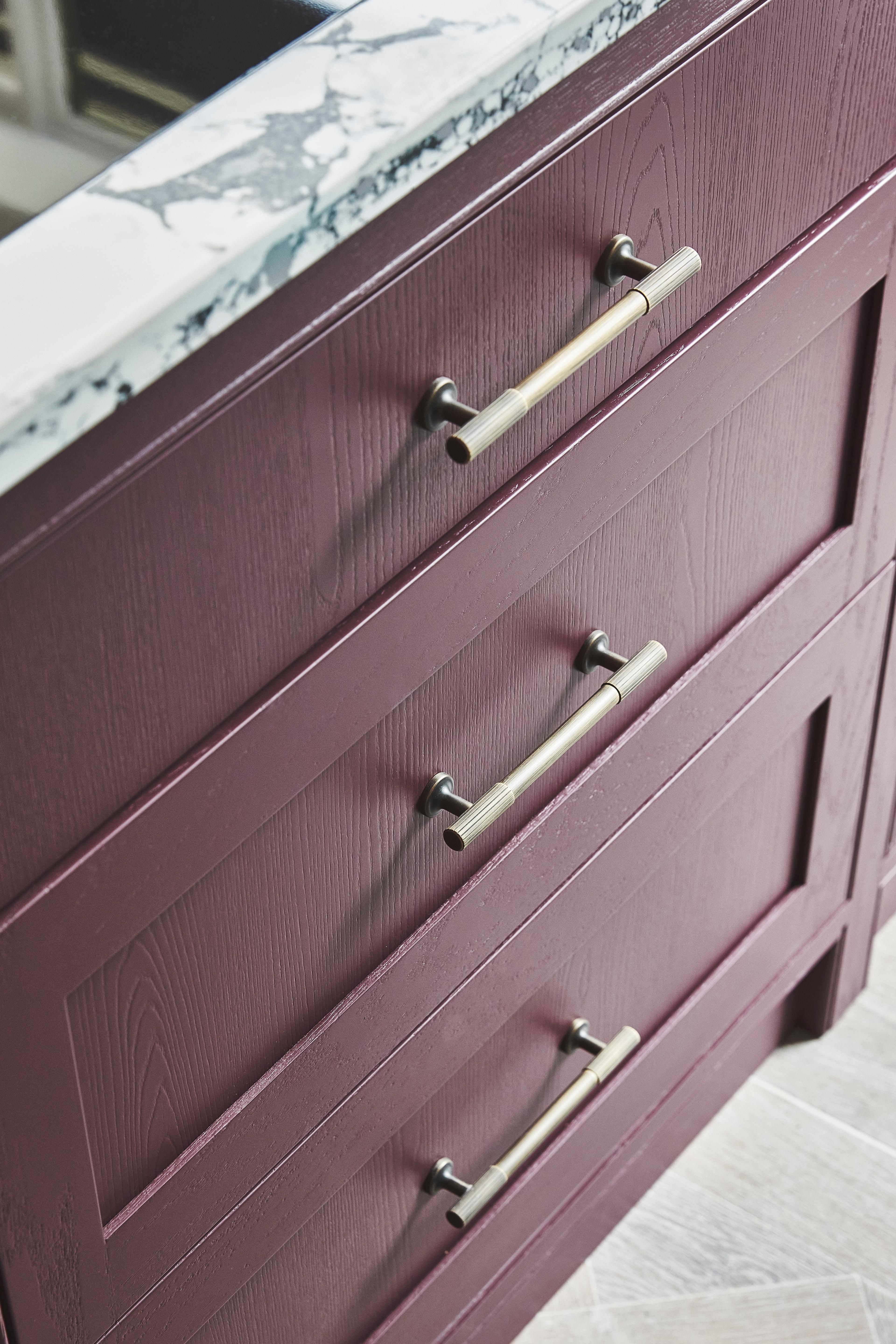 Close-up of a cabinet with three purple wooden drawers, each featuring a silver handle. The top is made of white marble with gray veining. The cabinet stands on a light-colored wooden floor.