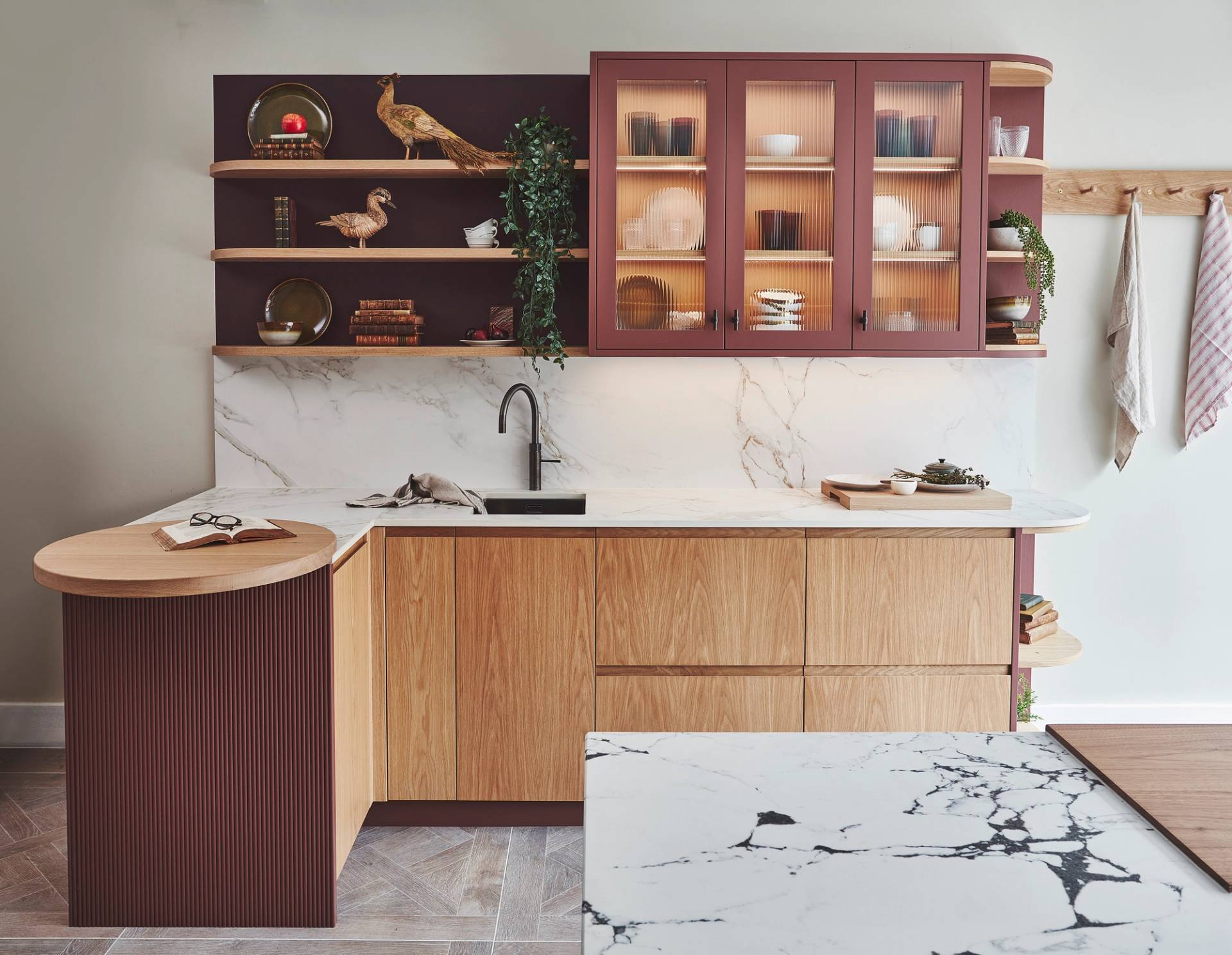 A contemporary kitchen interior showcases light wood cabinetry, burgundy fluted glass cupboards, and open wooden shelving set against a white marble backsplash.