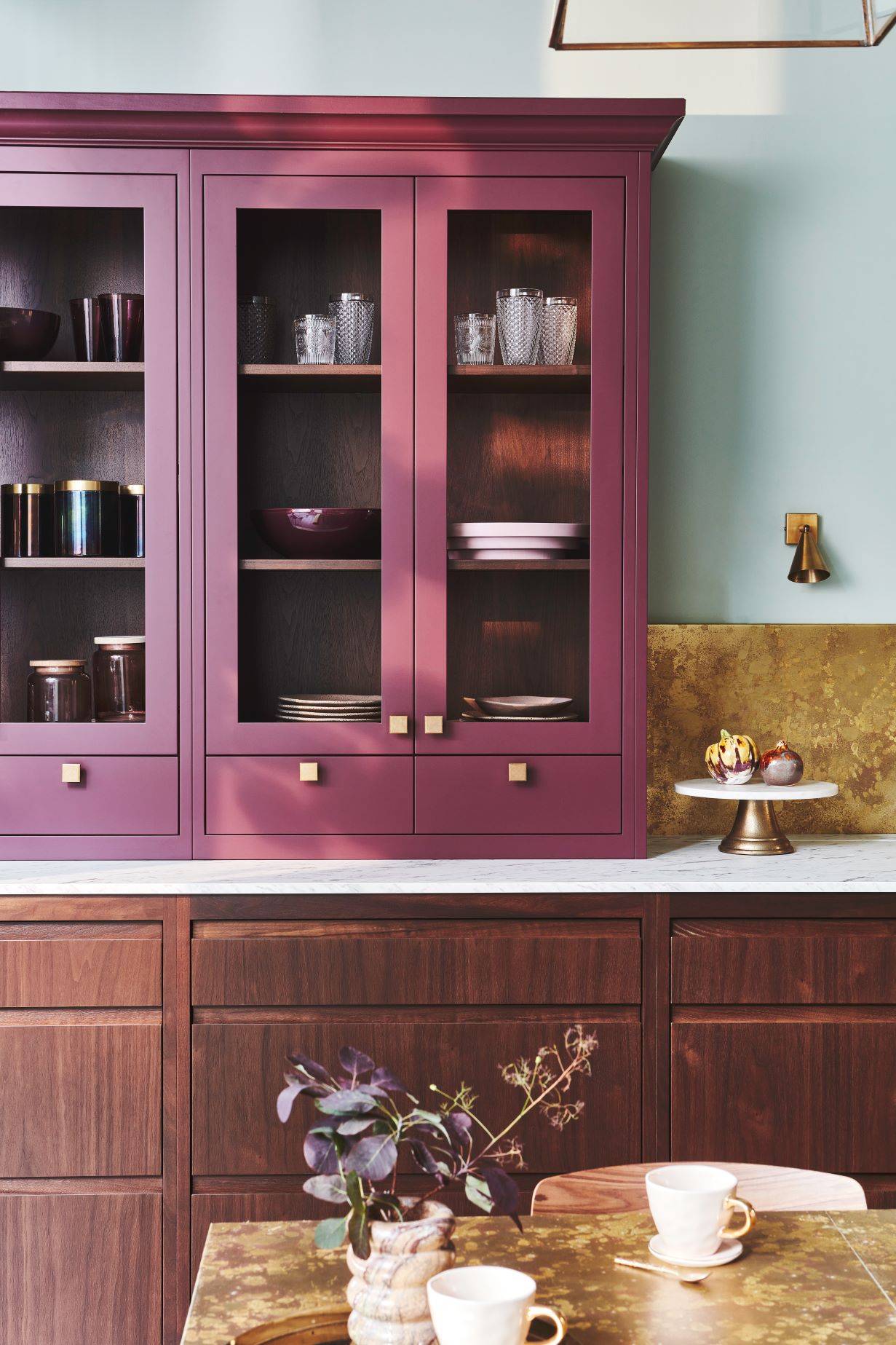 A kitchen with wooden cabinets and maroon accent cupboards with glass doors displaying dishes and glassware. The counter has a white marble top and a gold backsplash. A table in the foreground holds a plant, a cake stand with fruit, and two cups.