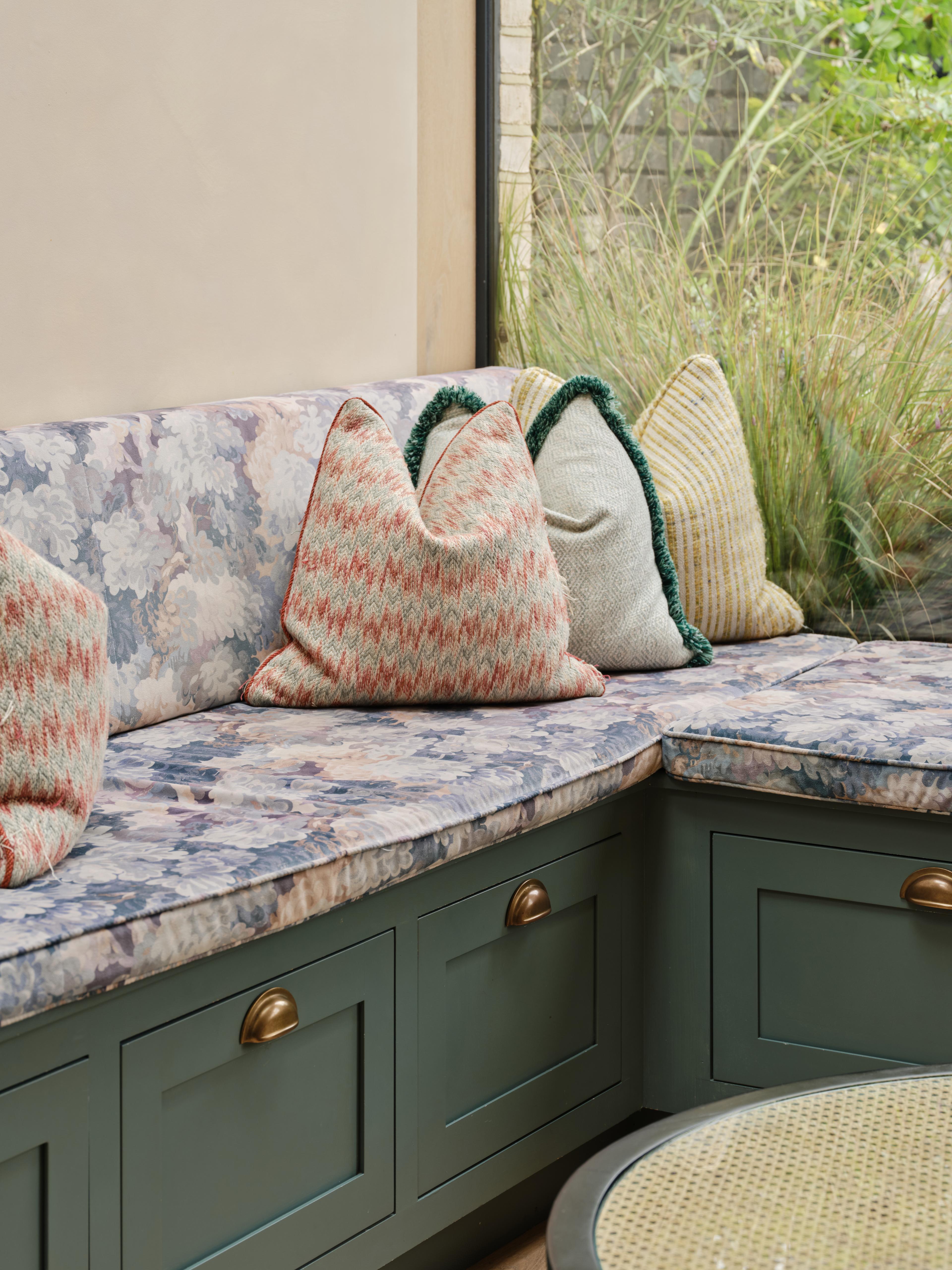 A cozy kitchen with a mix of styles. Light blue cabinets complement a marbled countertop and backsplash. A wooden shelf holds books and dishes, while a maroon island features a cutting board and red pot. Plants hang above the sink for decoration.
