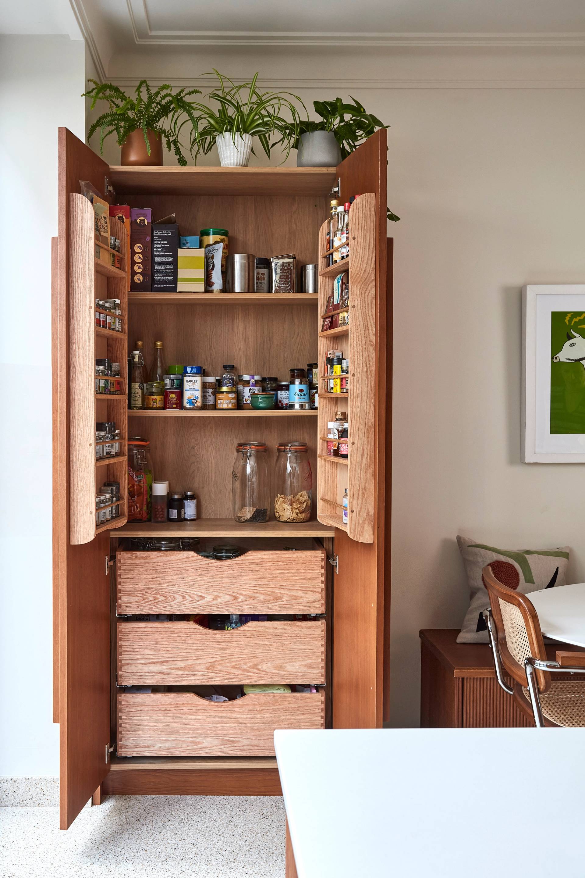 Tall kitchen cabinet with open doors filled with neatly organized spices, jars, and boxes. Two large glass jars are on a shelf, with plants on top of the cabinet. Next to the cabinet is a wooden chair and a framed wall art piece.
