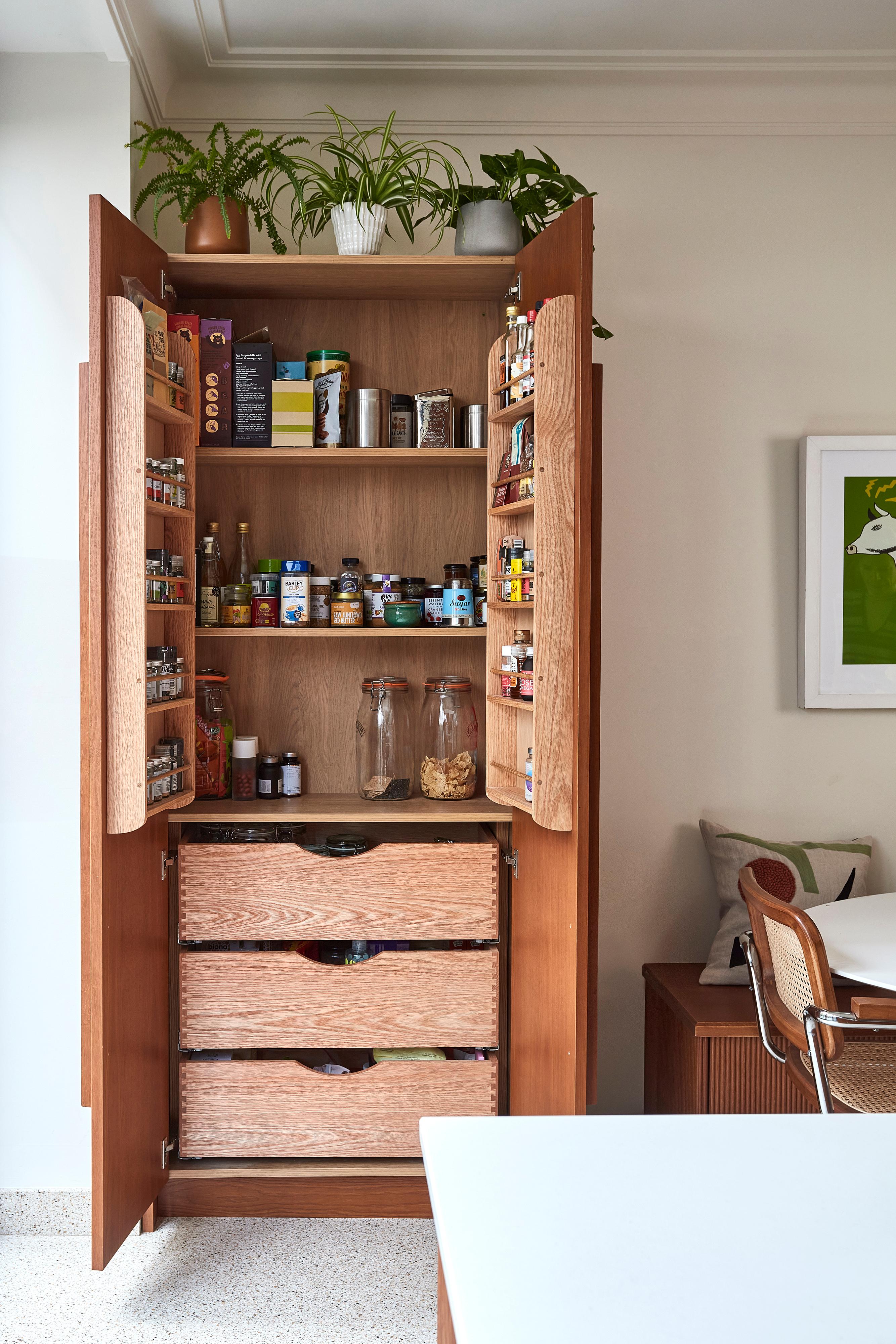 Tall kitchen cabinet with open doors filled with neatly organized spices, jars, and boxes. Two large glass jars are on a shelf, with plants on top of the cabinet. Next to the cabinet is a wooden chair and a framed wall art piece.