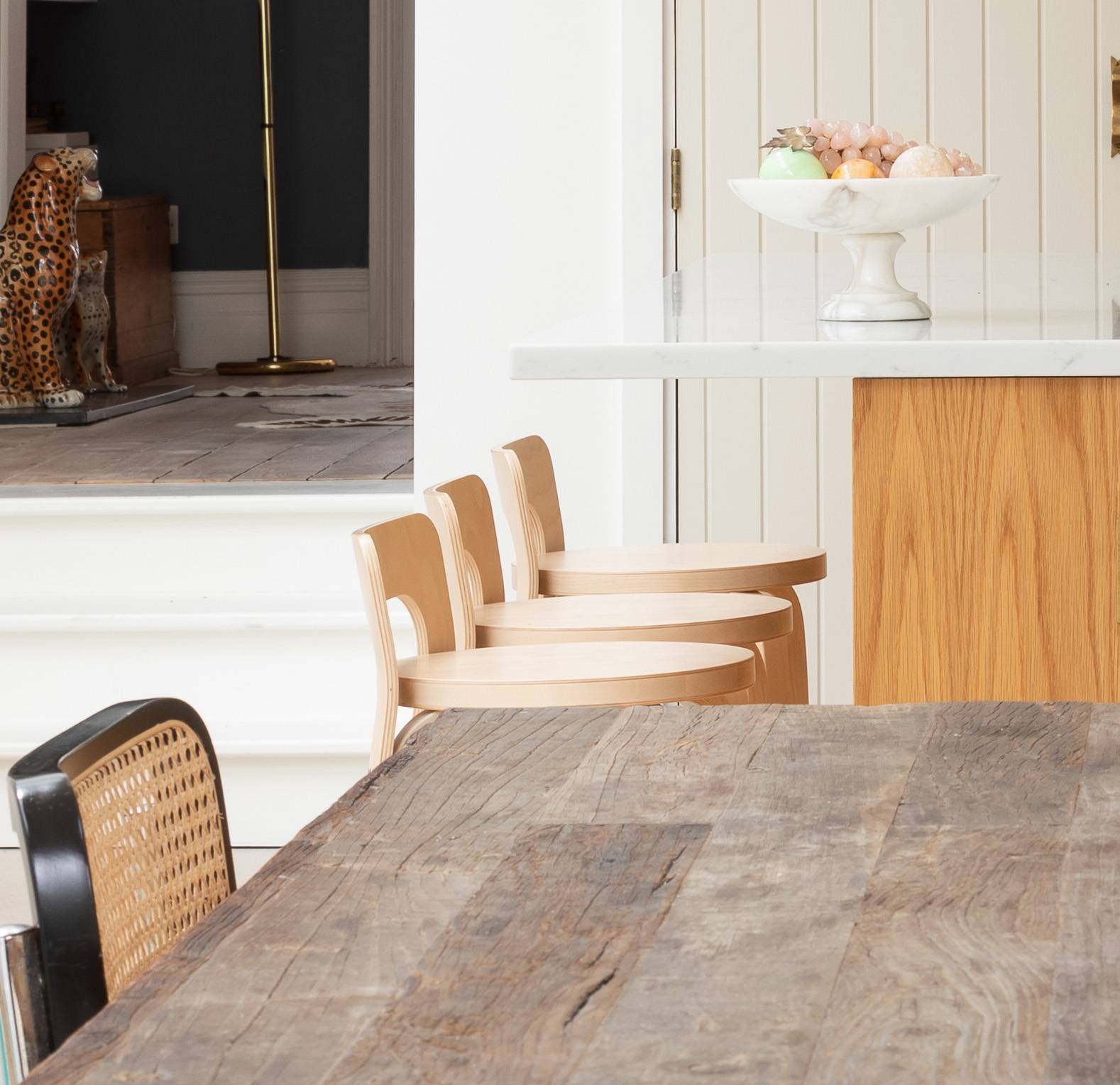 A rustic wooden dining table sits beside light wooden chairs and a kitchen island with a white countertop; a bowl of fruit is on the counter, and a decorative cheetah statue is visible in the background.