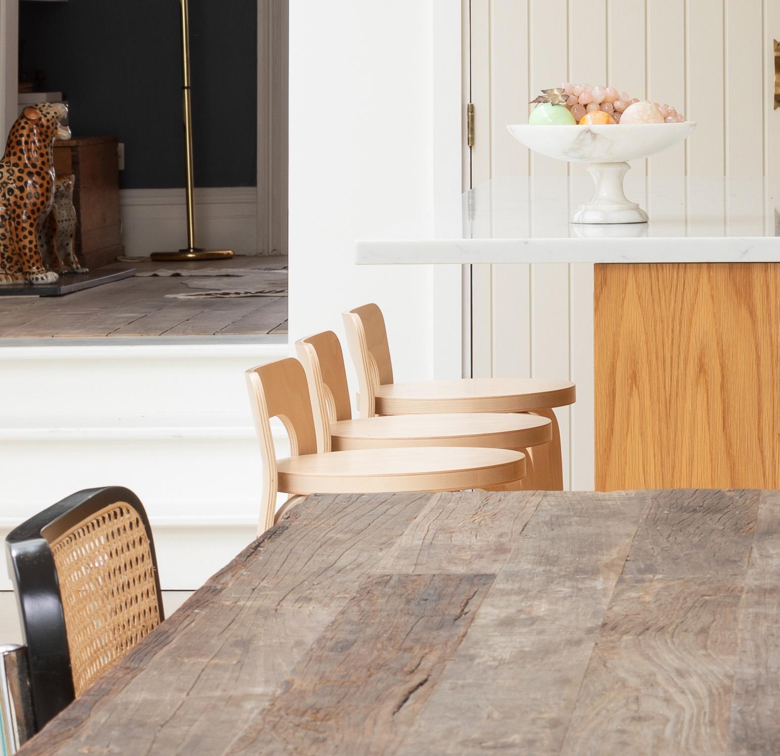 A rustic wooden dining table sits beside light wooden chairs and a kitchen island with a white countertop; a bowl of fruit is on the counter, and a decorative cheetah statue is visible in the background.