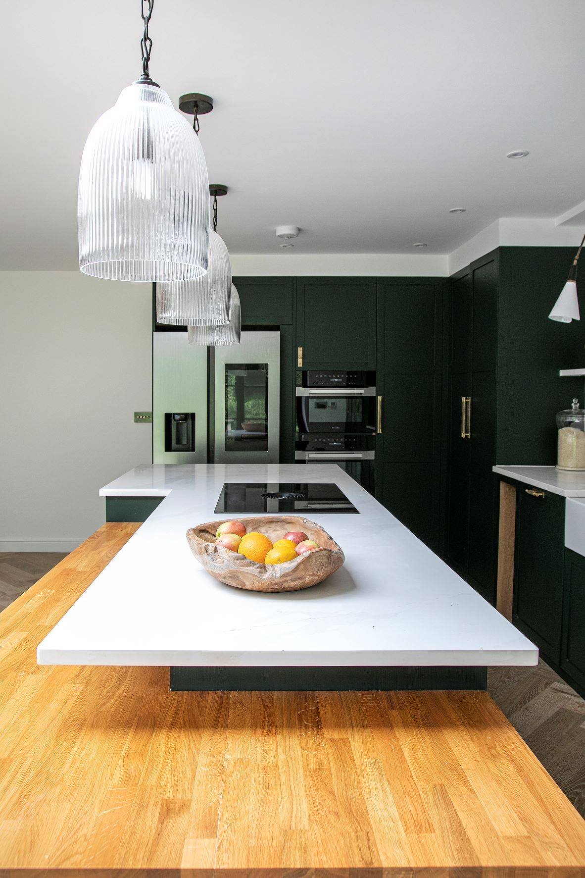 Modern kitchen with dark green cabinets and a large white kitchen island with a wooden extension. Three pendant lights hang above the island. A wooden bowl with fruit sits on the counter. Stainless steel fridge and built-in oven are visible
