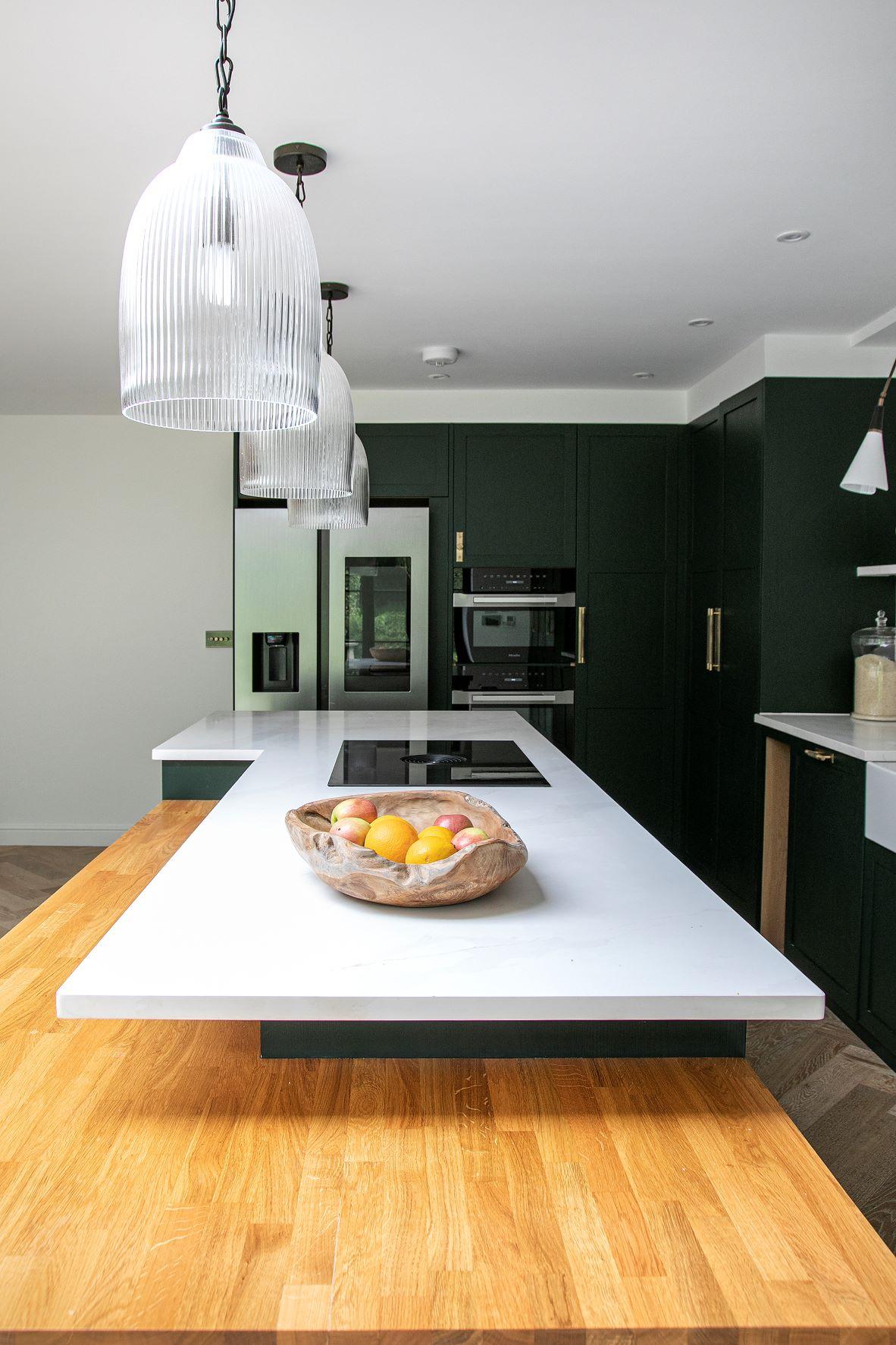 Modern kitchen with dark green cabinets and a large white kitchen island with a wooden extension. Three pendant lights hang above the island. A wooden bowl with fruit sits on the counter. Stainless steel fridge and built-in oven are visible