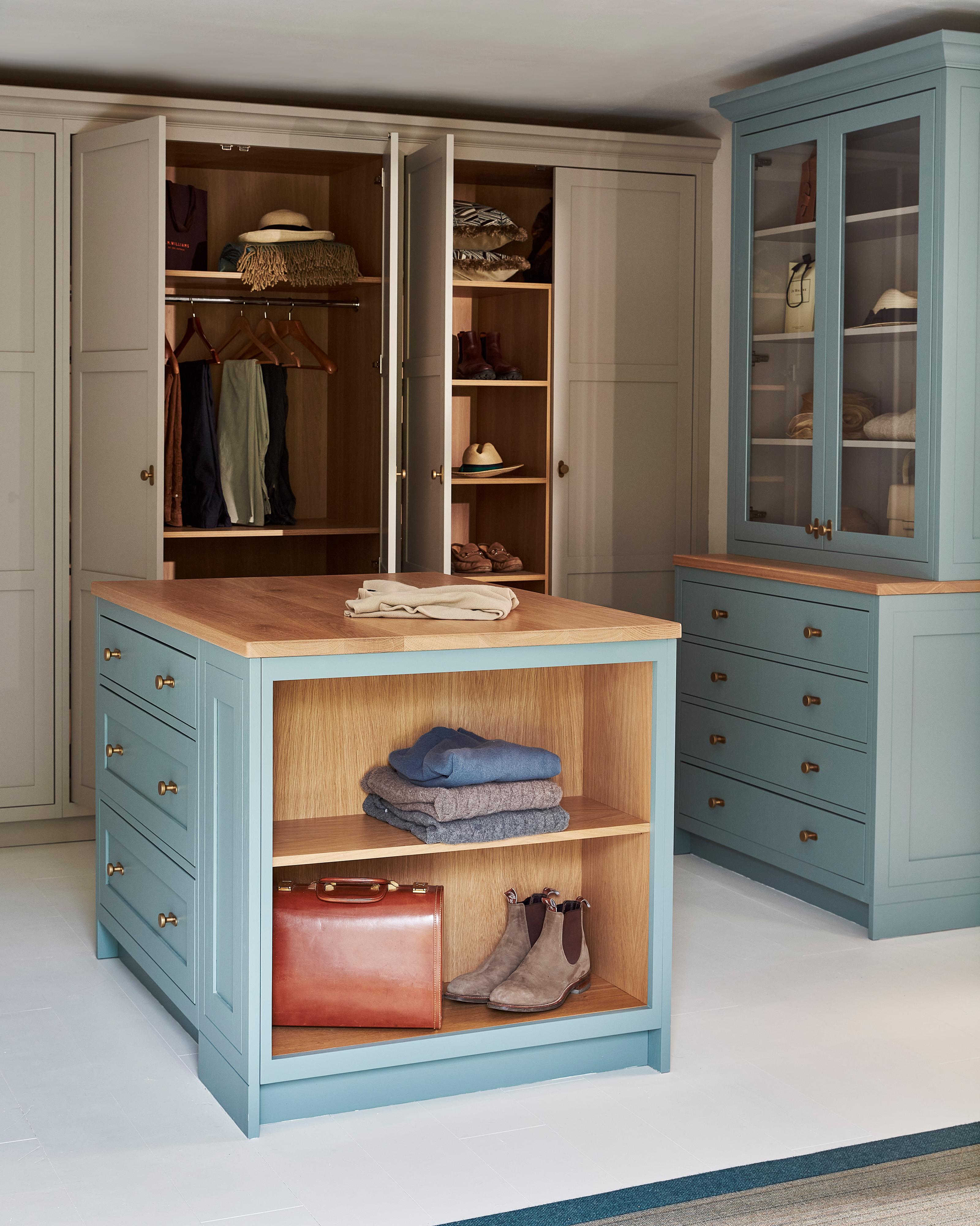 A spacious walk-in closet with blue cabinetry, open shelves displaying folded clothes, boots, and a leather bag. An island with drawers is in the center, and open wardrobes reveal hanging clothes and hats.