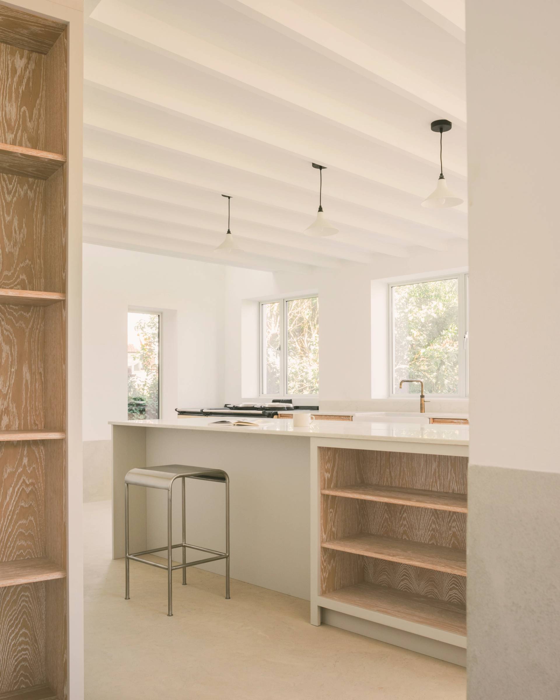 a bright, modern kitchen features a central island with a white countertop and built-in wooden shelving, a metal bar stool, and three pendant lights hanging from a white beamed ceiling.