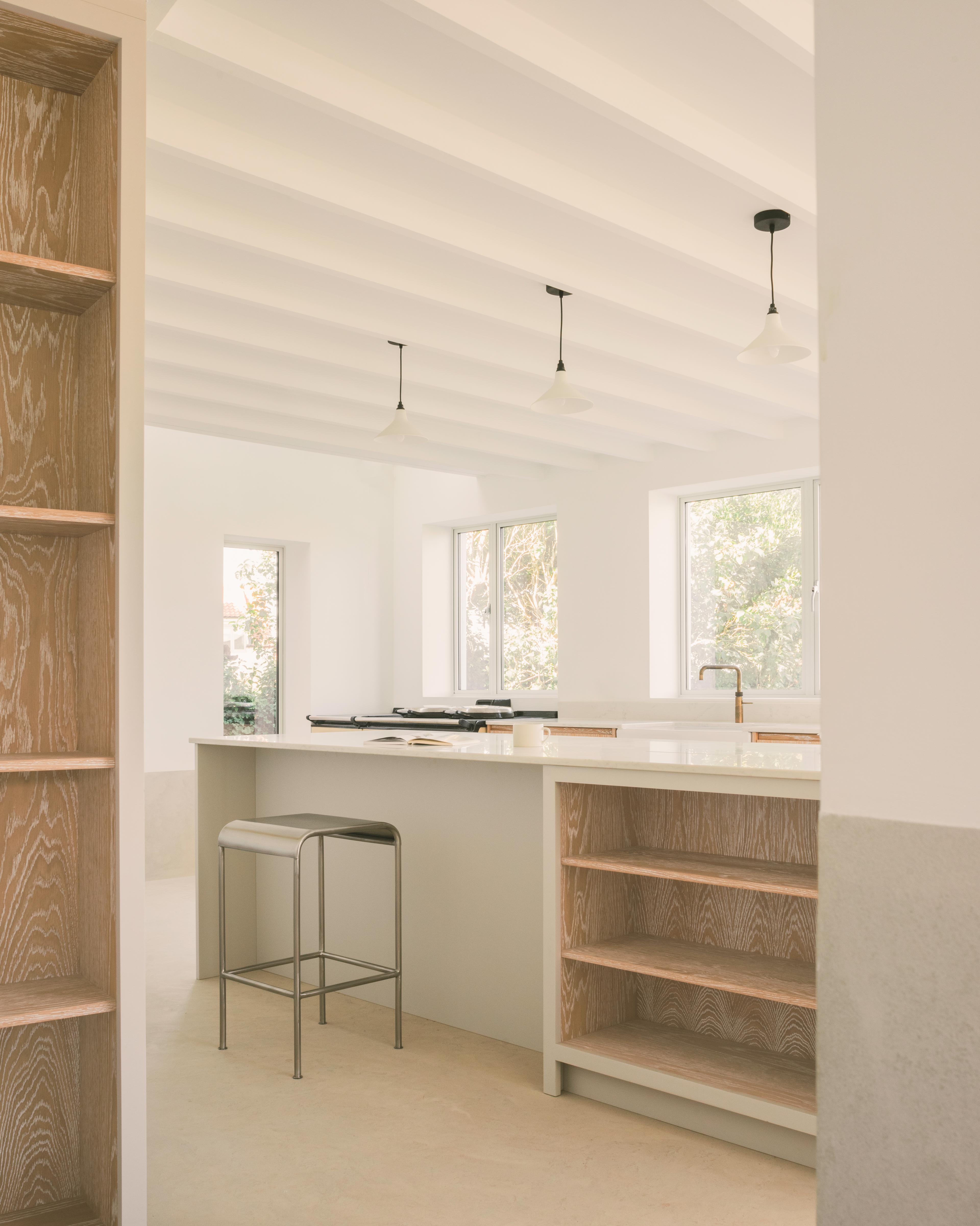 a bright, modern kitchen features a central island with a white countertop and built-in wooden shelving, a metal bar stool, and three pendant lights hanging from a white beamed ceiling.