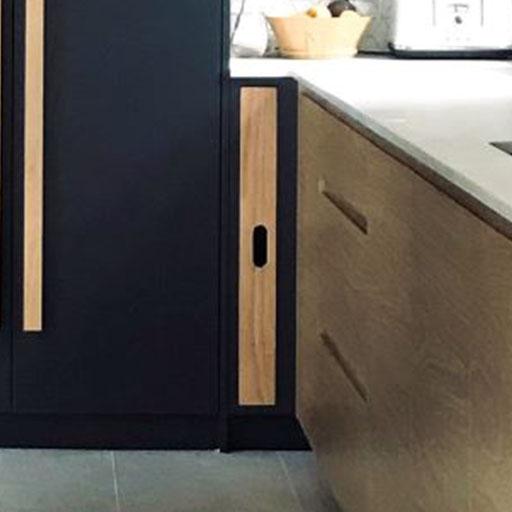 A small, narrow cabinet with a wood finish is built into a kitchen, positioned between a tall black cabinet and a light wood counter with drawers.