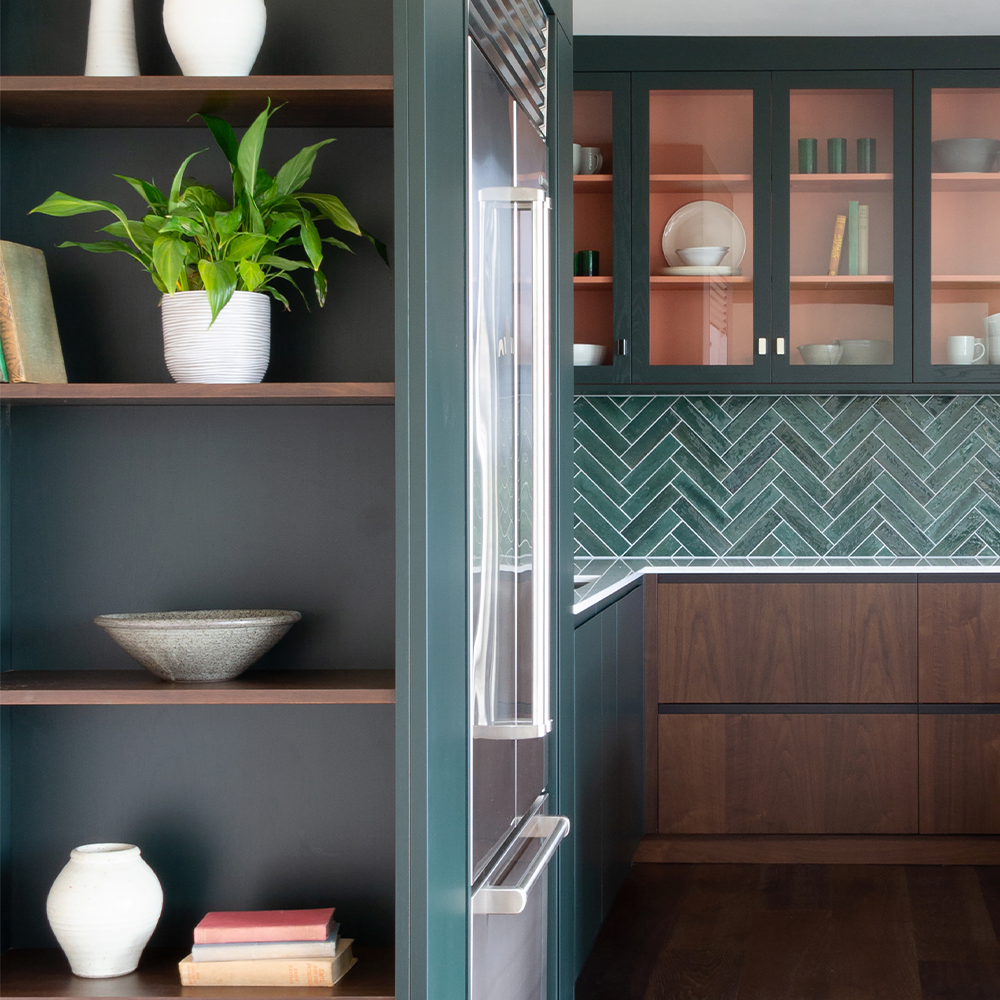 A modern kitchen with dark cabinetry, open shelves holding books and ceramics, a potted plant, green herringbone tile backsplash, and glass-front upper cabinets displaying neatly arranged dishes.