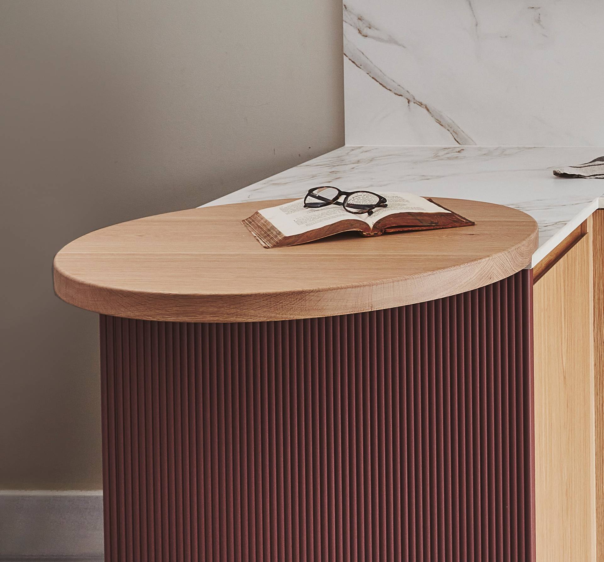 A round wooden side table with a closed book and a pair of glasses on top. The table has a dark, ribbed base. In the background is a marble countertop and a beige wall.
