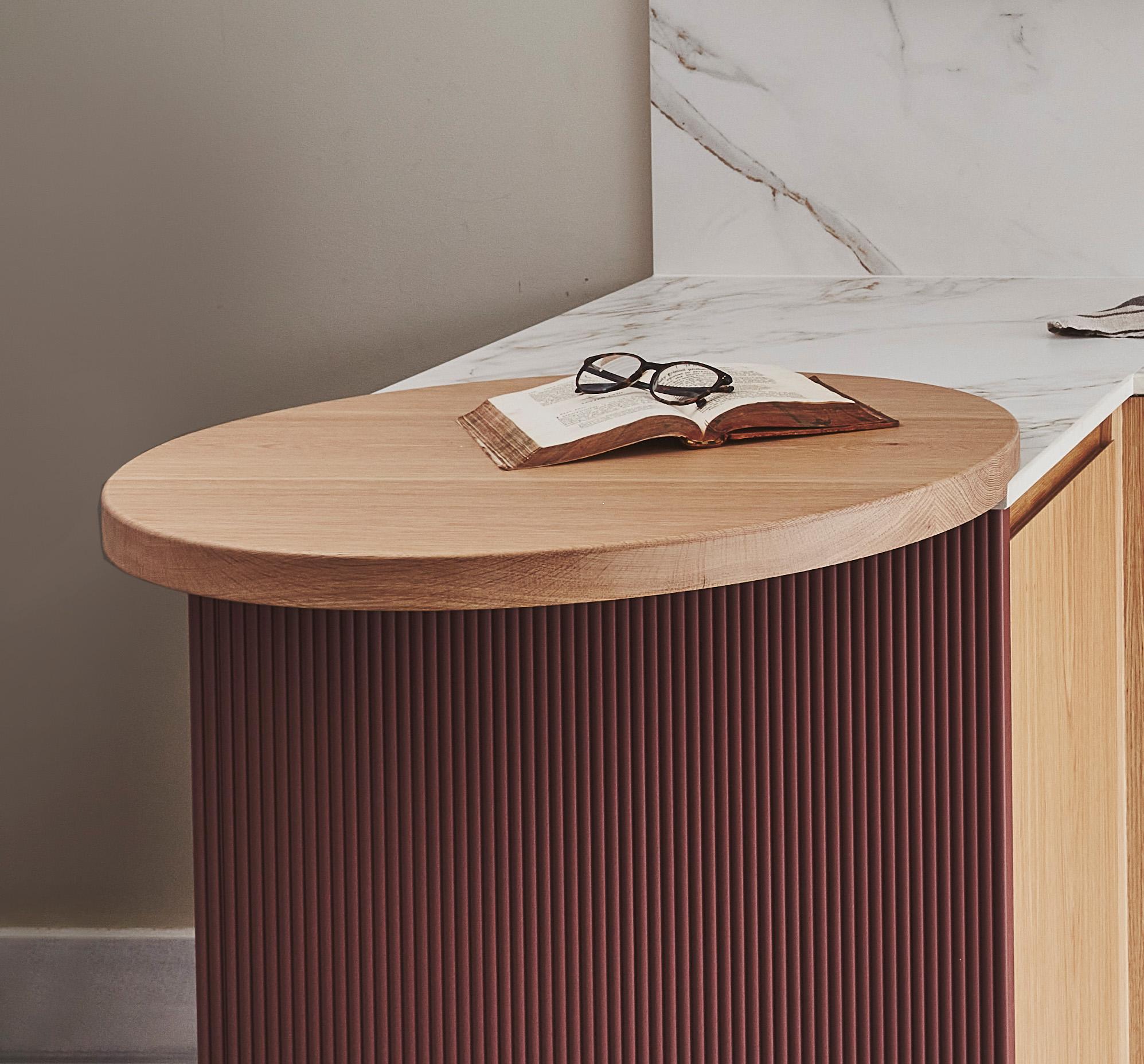 A round wooden side table with a closed book and a pair of glasses on top. The table has a dark, ribbed base. In the background is a marble countertop and a beige wall.
